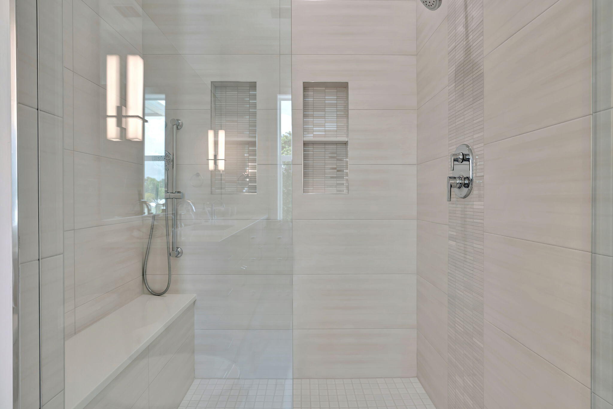 Walk-in shower with clear glass, pale tile, recessed niches, a bench, and chrome fixtures.