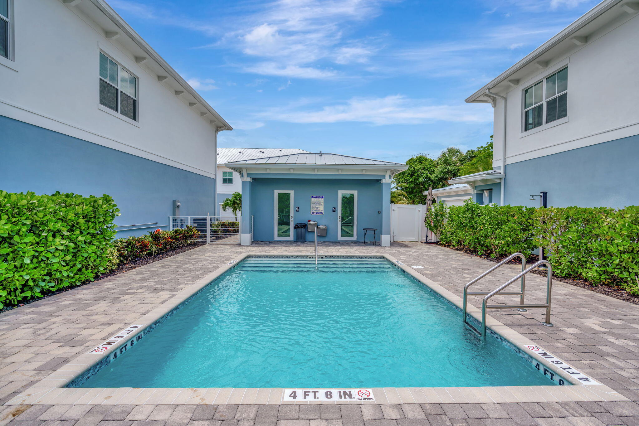 Outdoor swimming pool between blue-and-white buildings, with a paved deck and metal ladder.