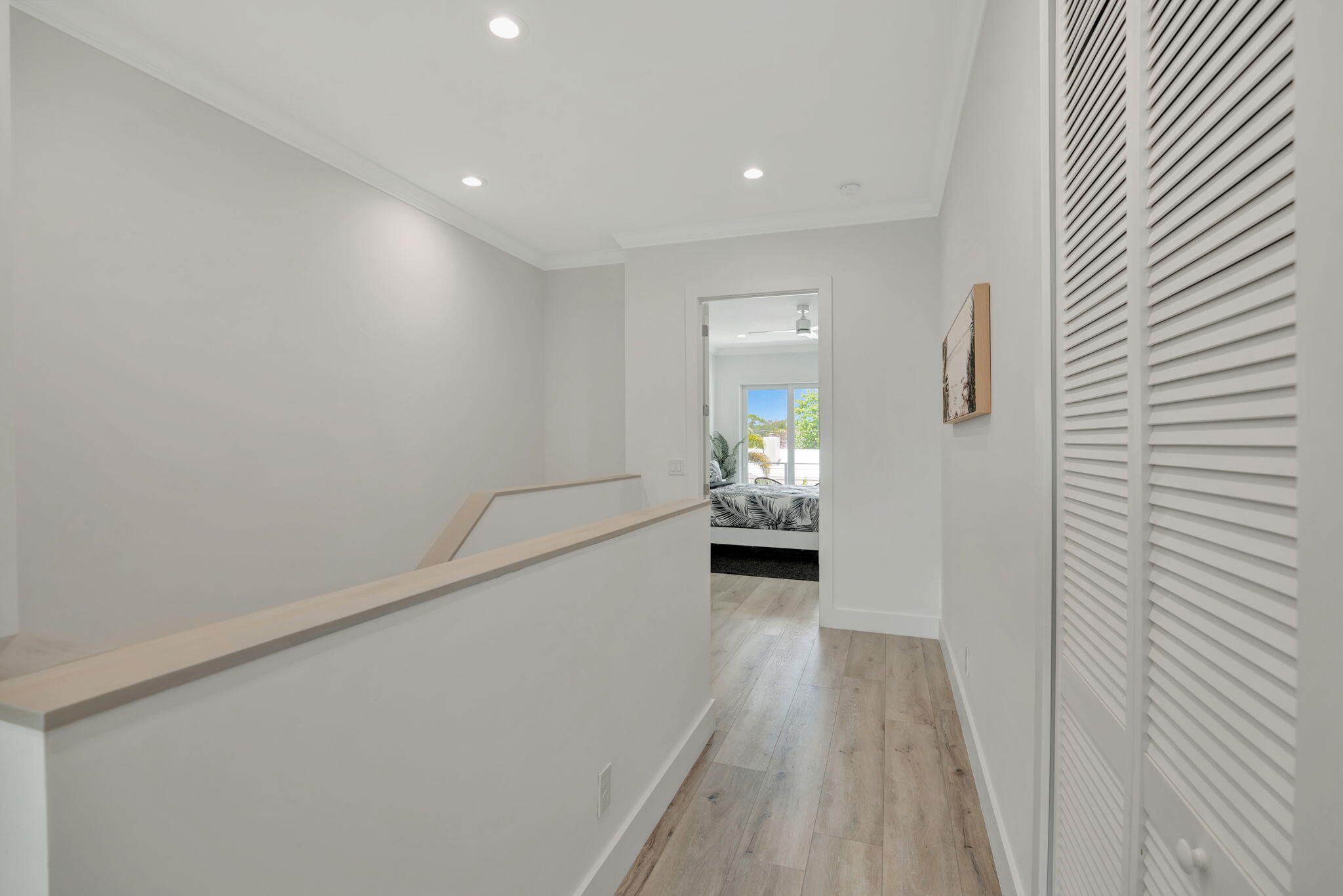 Upstairs hallway with pale wood flooring and louvered closet doors, opening onto a furnished bedroom.