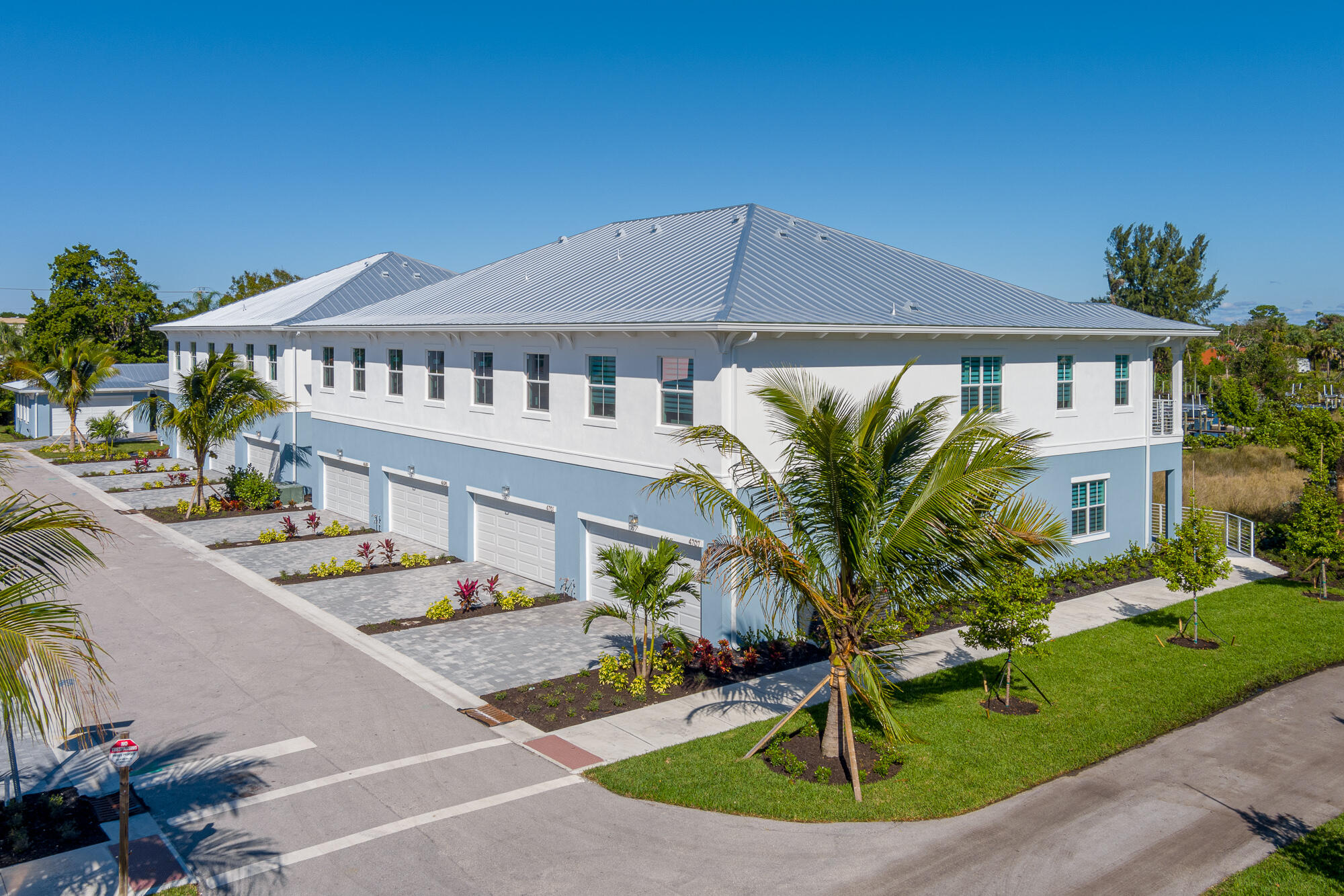 Two-story white and blue building with a silver roof, multiple garages, palm trees, and marina nearby.