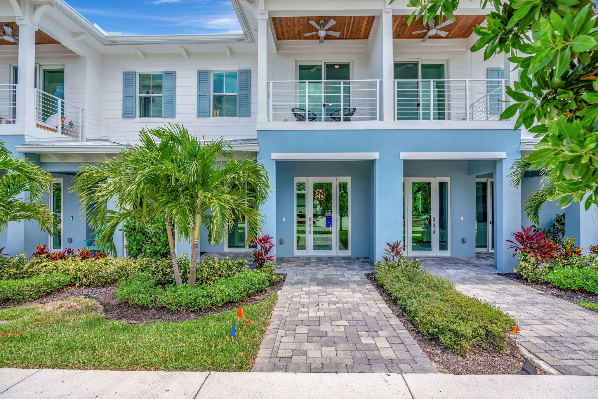 Two-story white and blue home with covered balconies, tropical landscaping, and a paved front walkway.