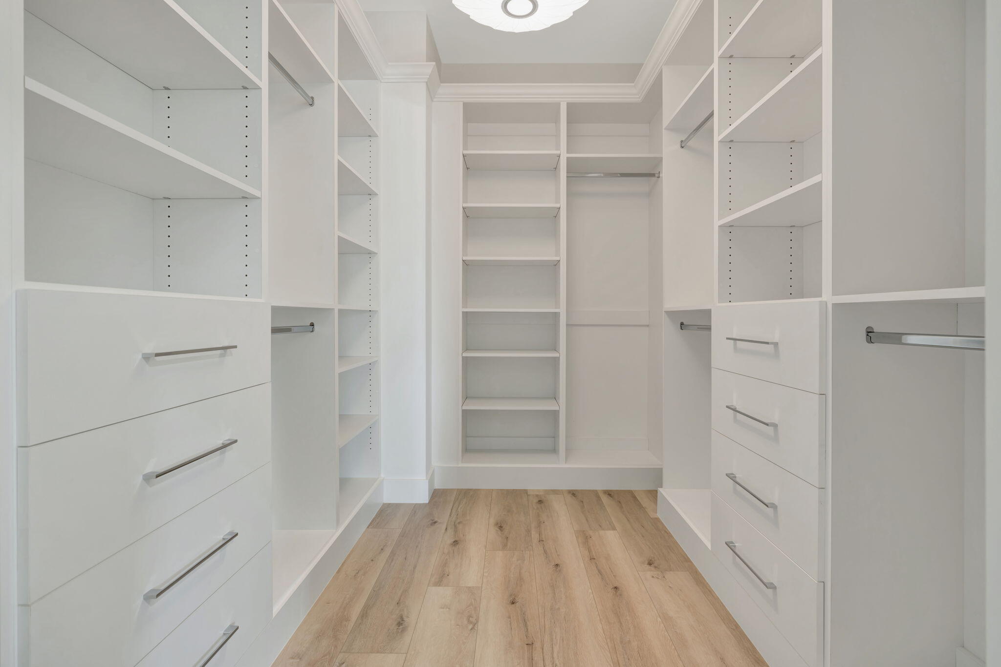 Walk-in closet with white built-in shelving and drawers, hanging rods, and light wood flooring.