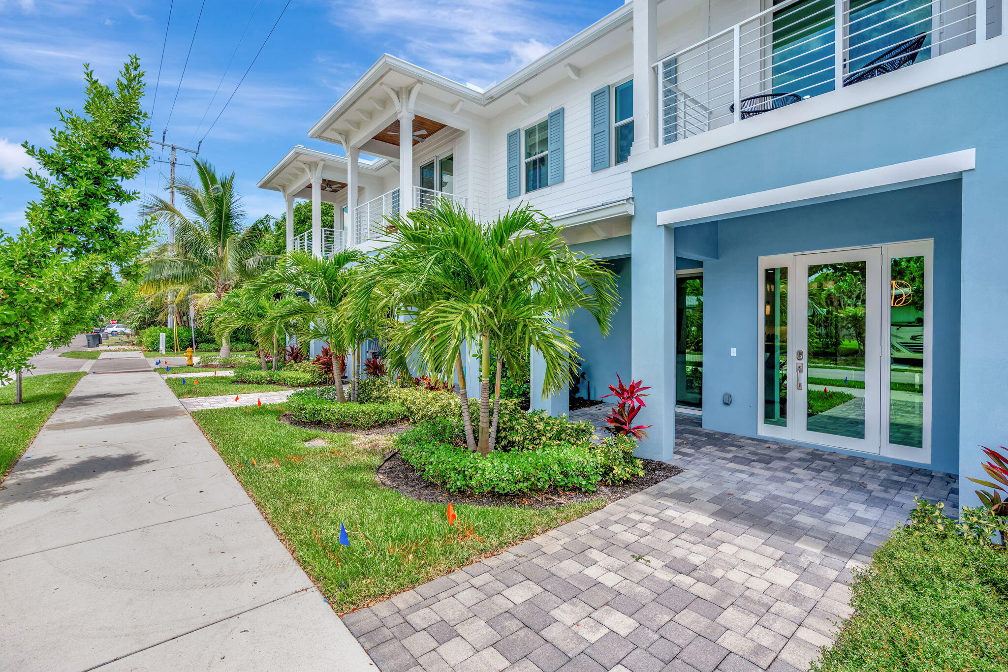 Two-story white and blue home with tropical landscaping, glass entry doors, and upper balconies.