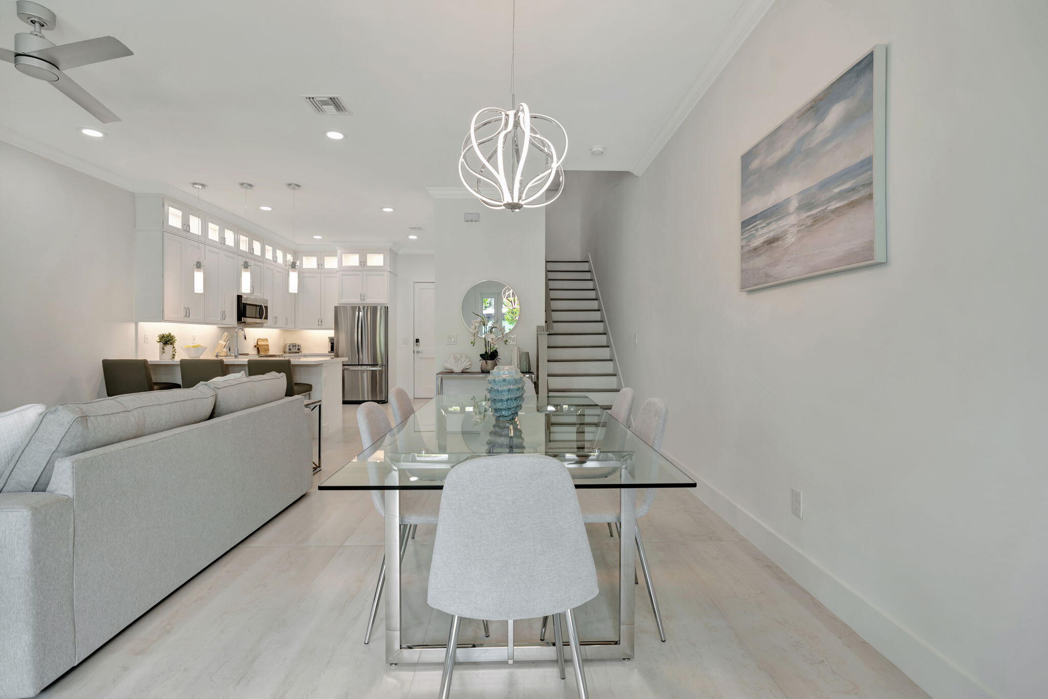 Open-plan dining and living area with a glass table, gray sofa, white kitchen, and staircase.