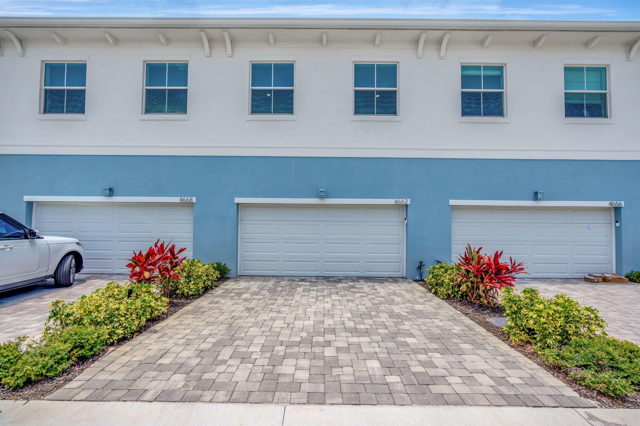 Blue-and-white building exterior with three garage doors, paver driveway, and tropical landscaping.