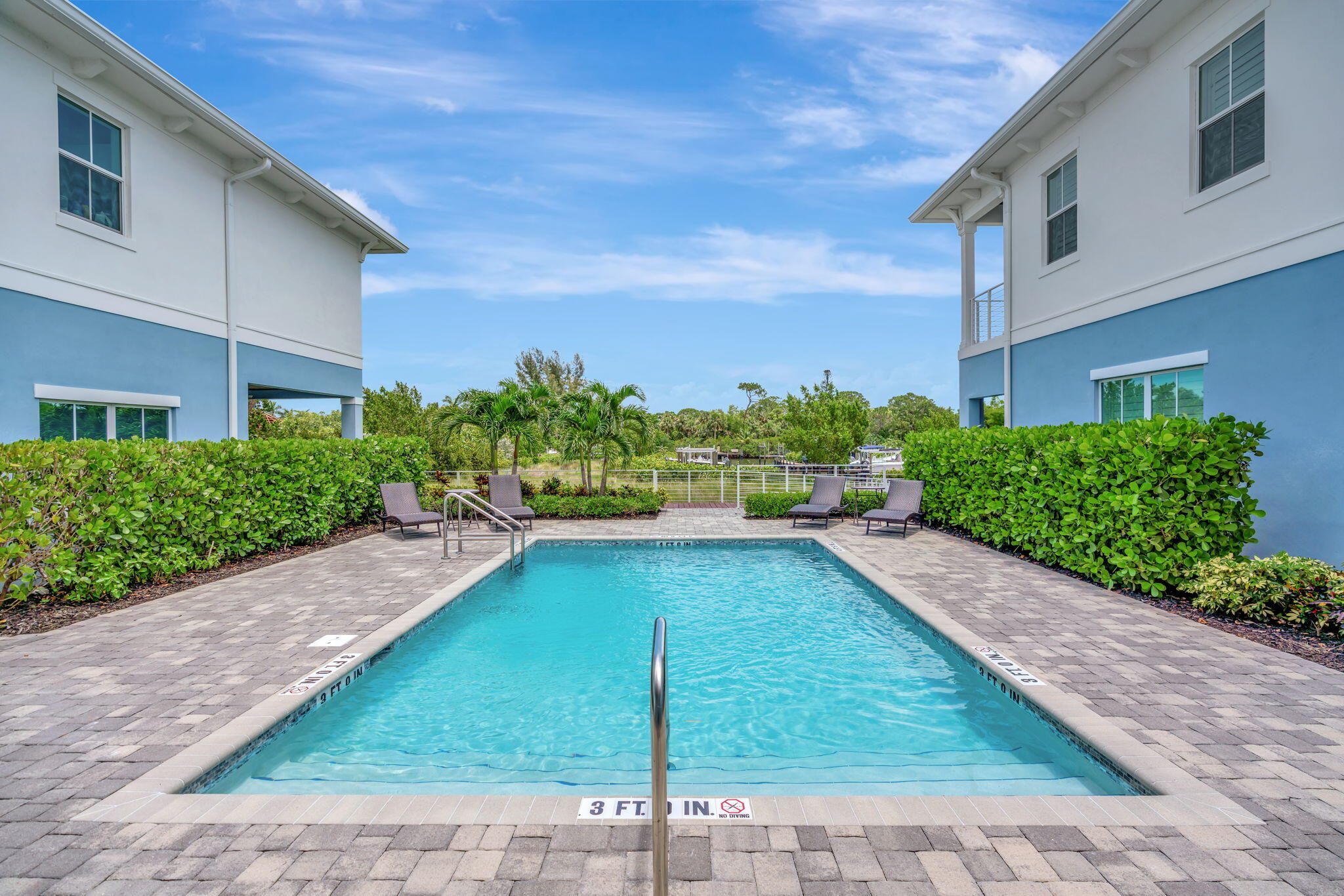 Outdoor pool between blue-and-white buildings, with a paved deck, lounge chairs, and palm trees.
