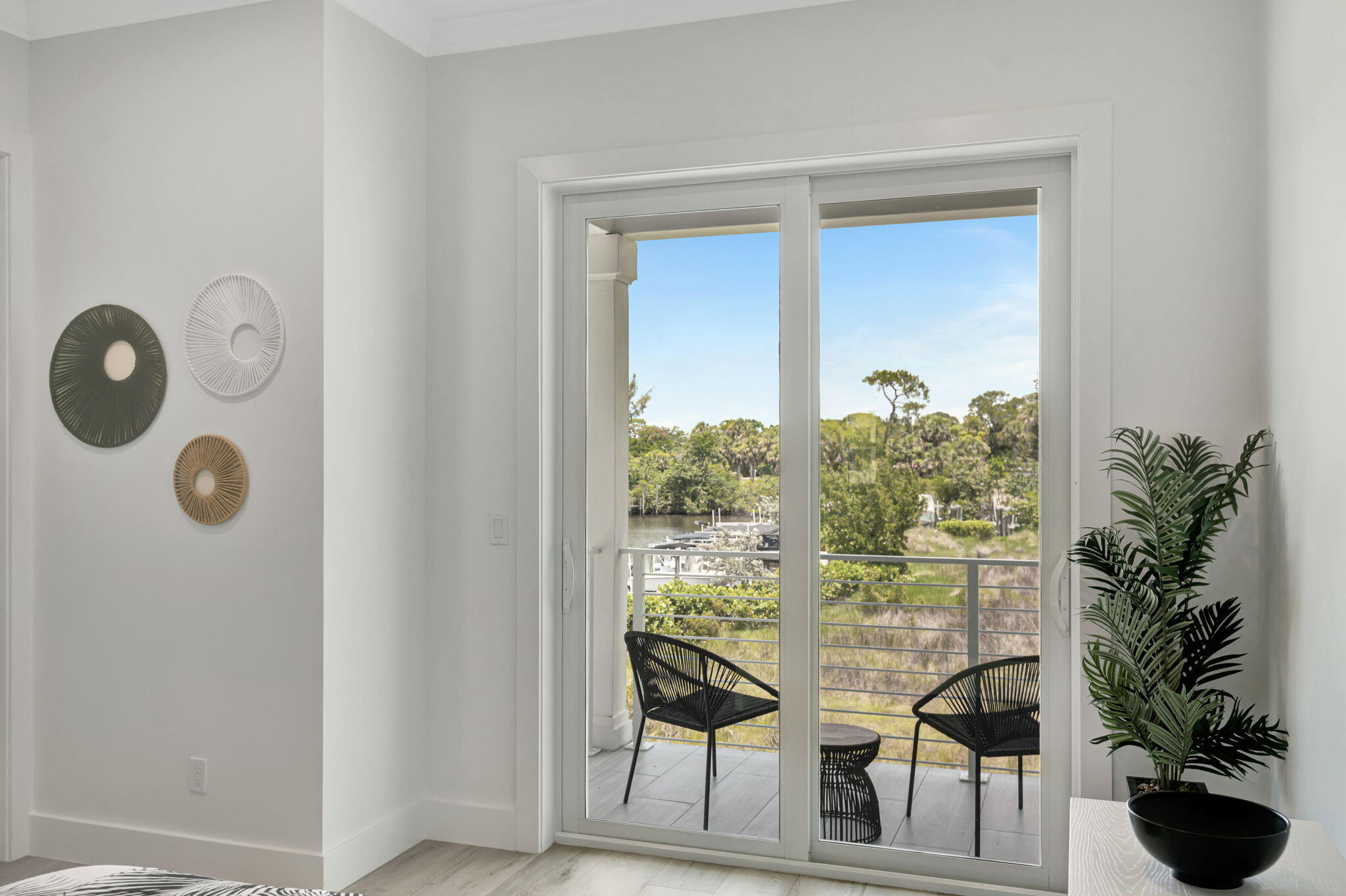 Bright interior with a sliding glass door opening to a furnished balcony overlooking water and trees.