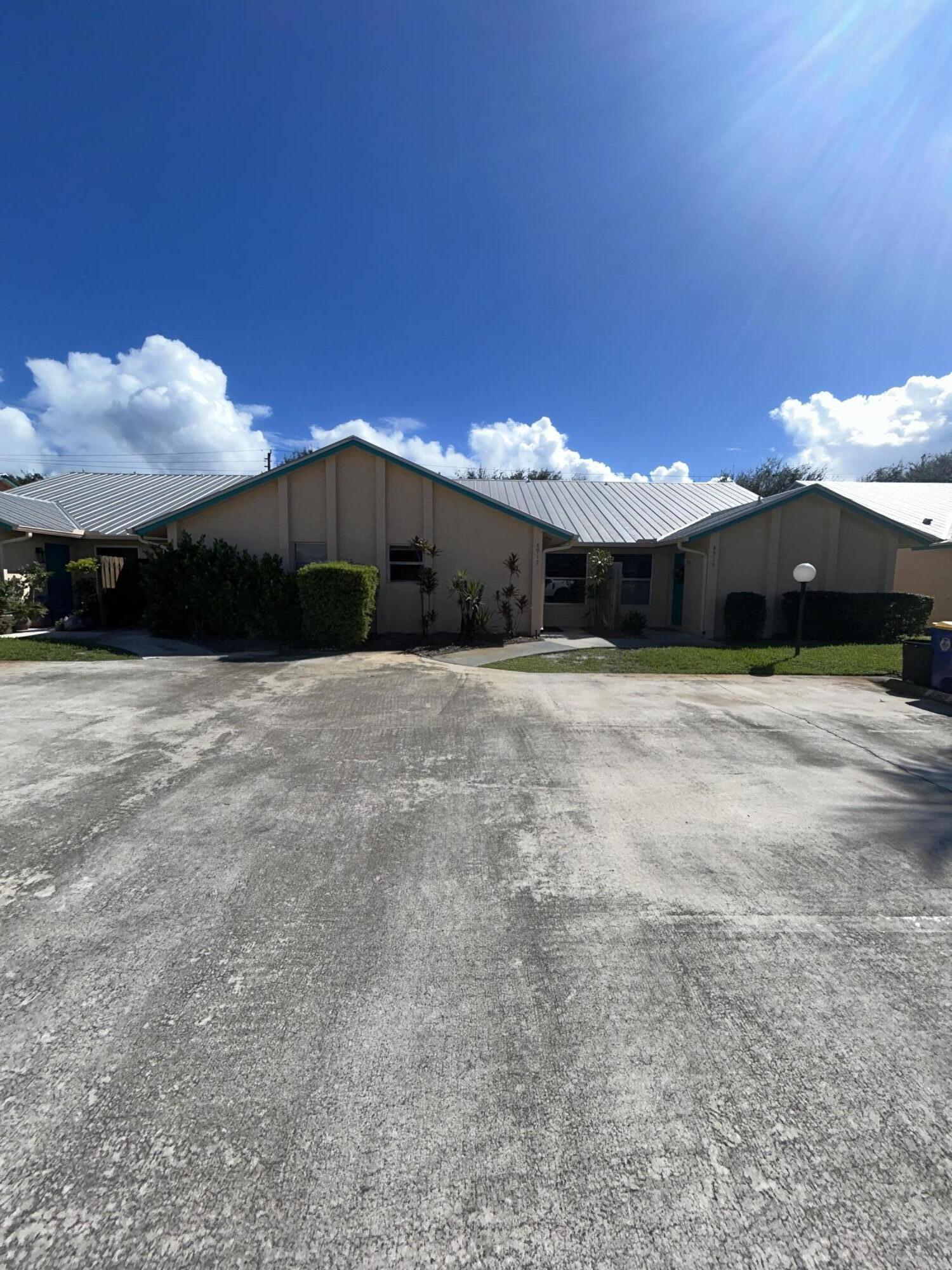 Beige low-rise property with a light metal roof, front shrubs, and a broad paved driveway.