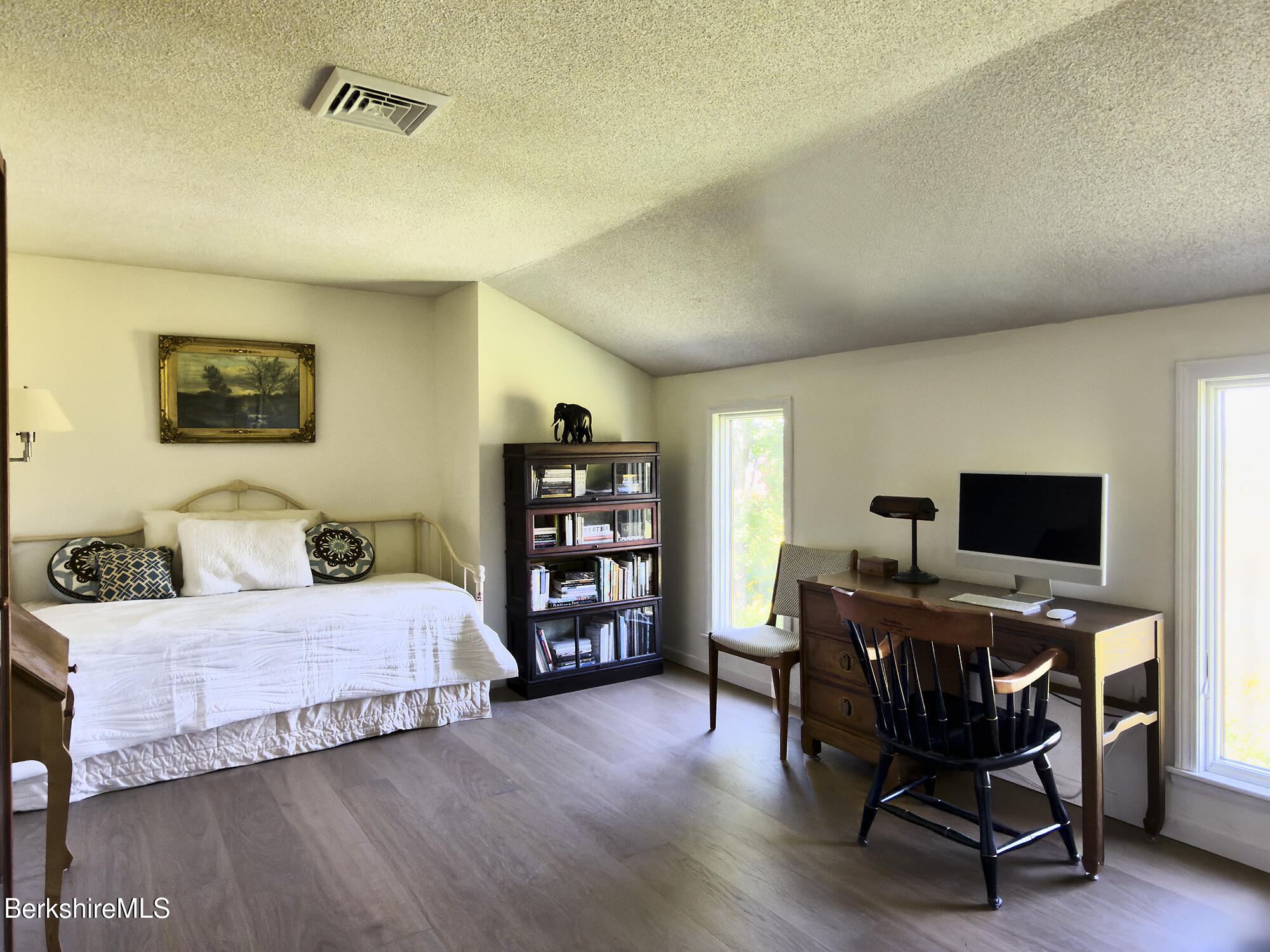 Bedroom with a white-covered bed, bookcase, and wooden desk beside tall windows.