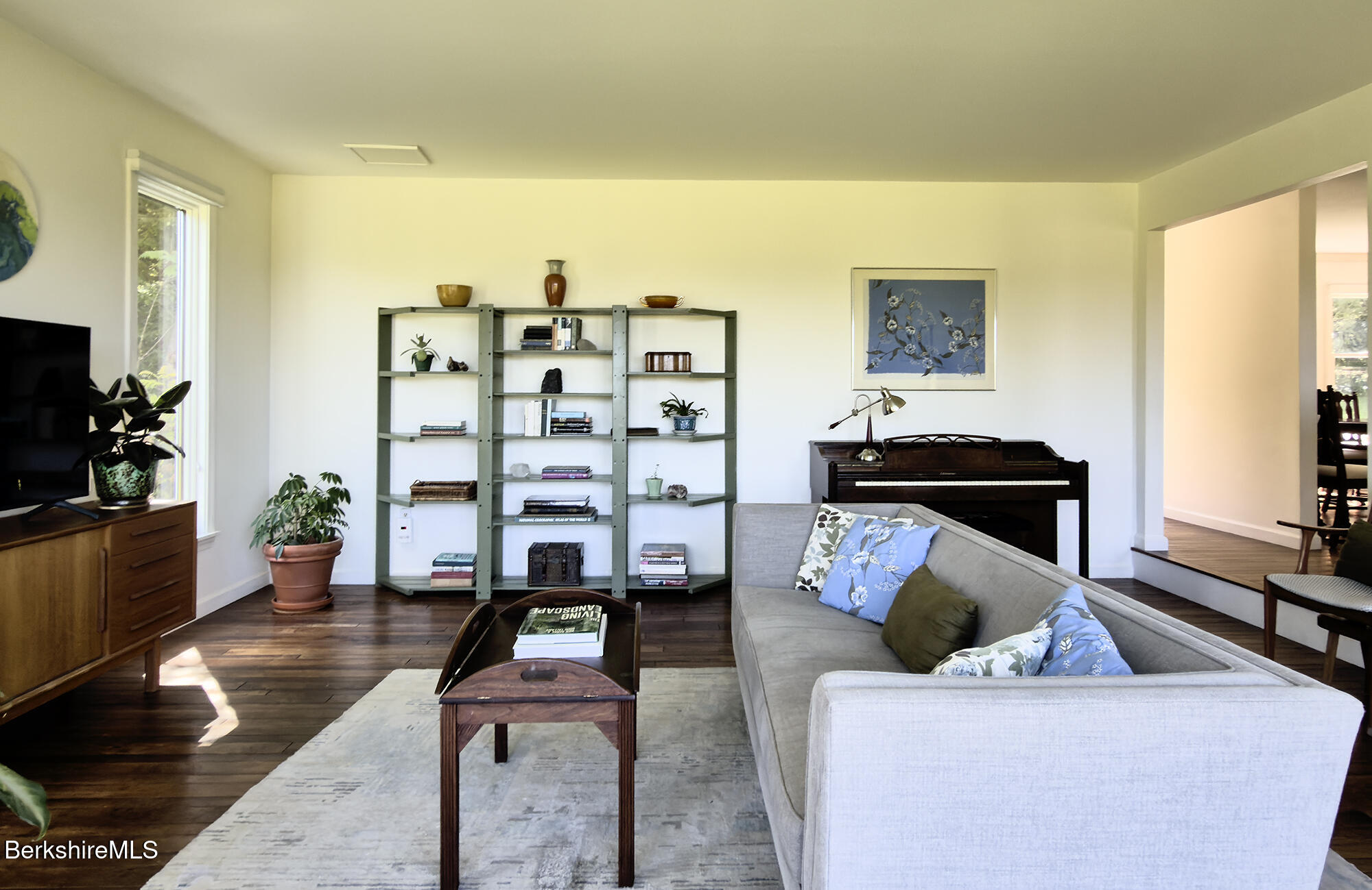 Living room with a gray sectional, dark hardwood floors, open shelving, and a piano.