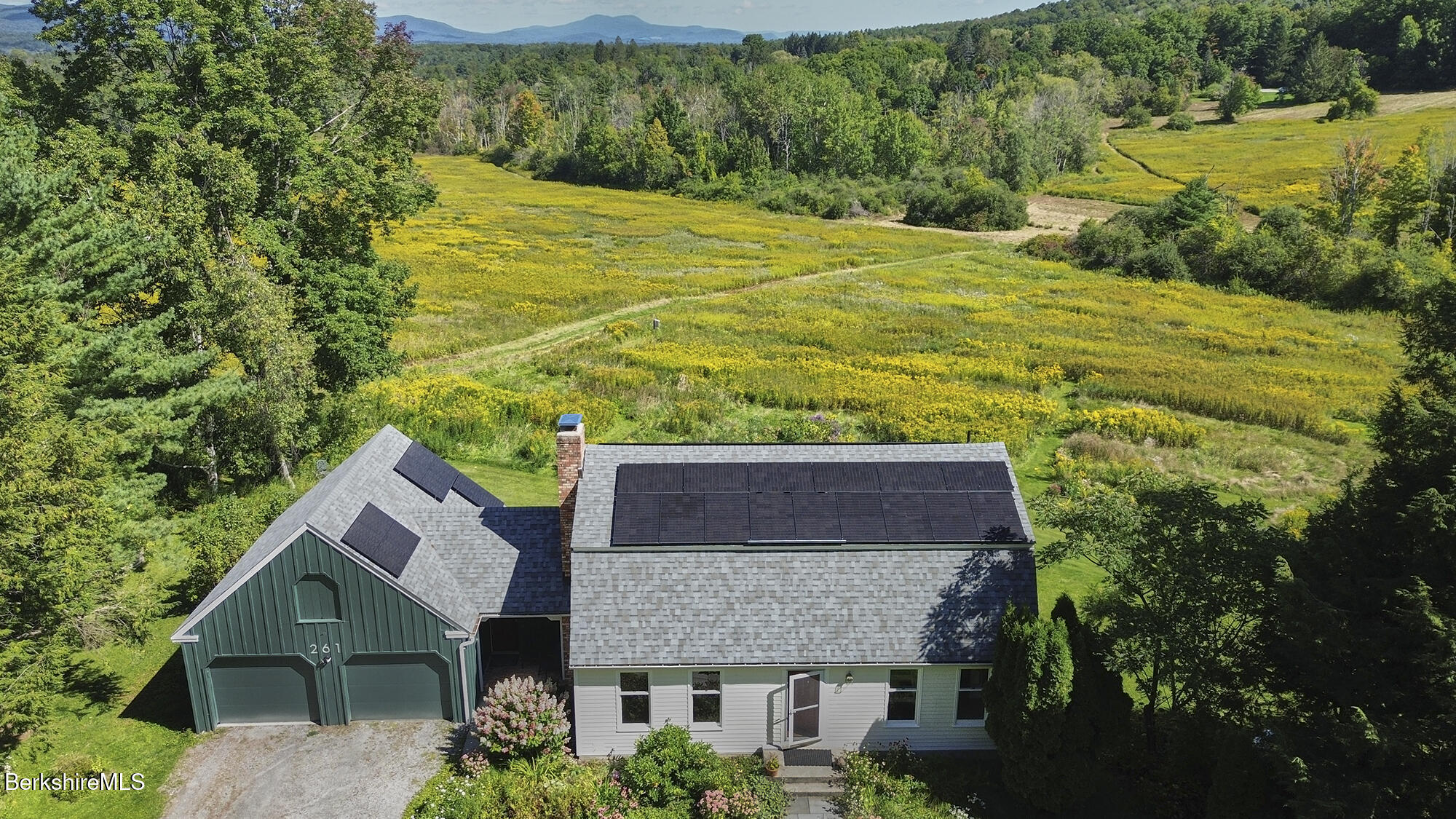 Aerial view of a house with rooftop solar panels beside a broad meadow and wooded hills.