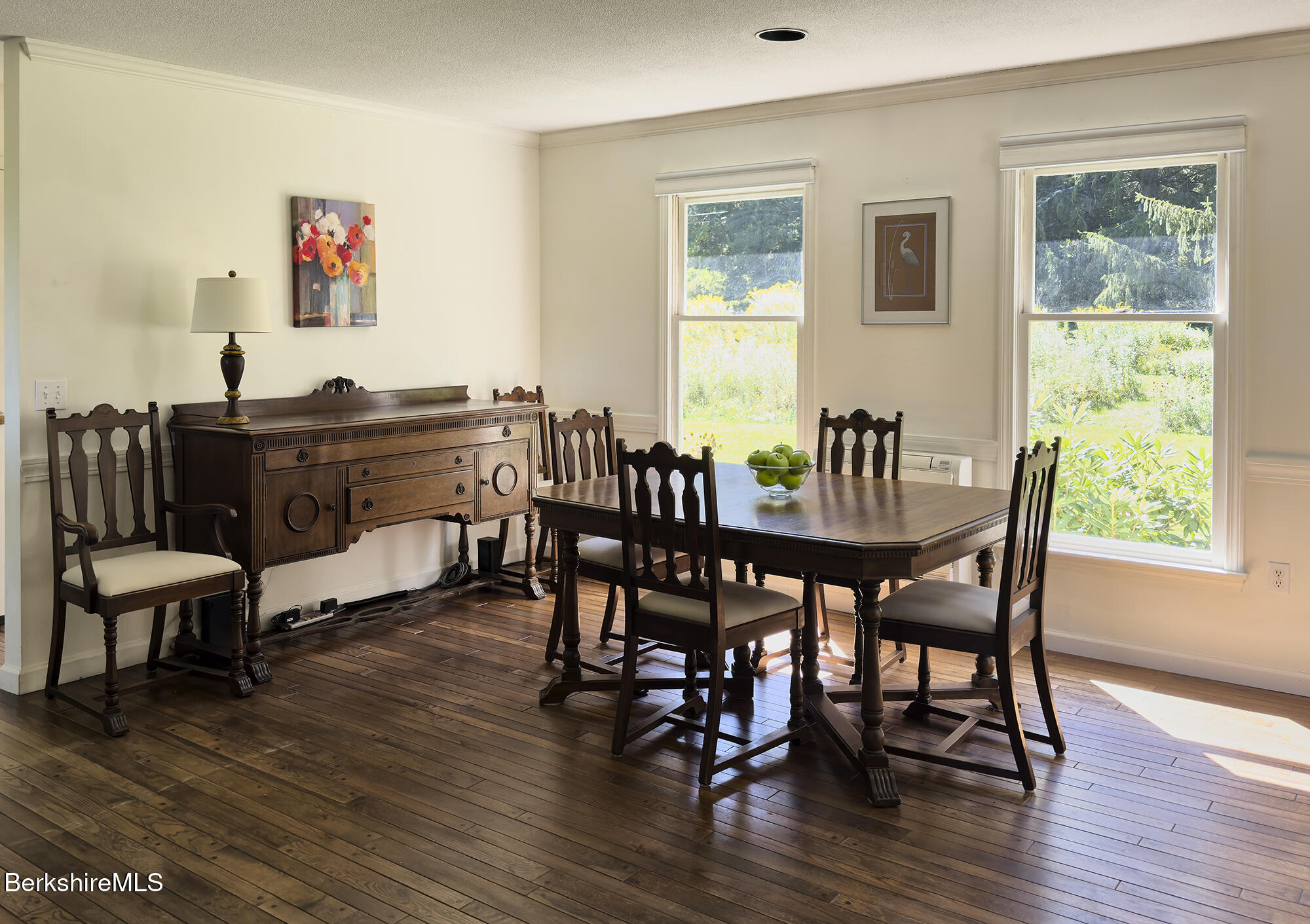 Dining room with a dark wood table and sideboard, hardwood floors, and large windows overlooking greenery.