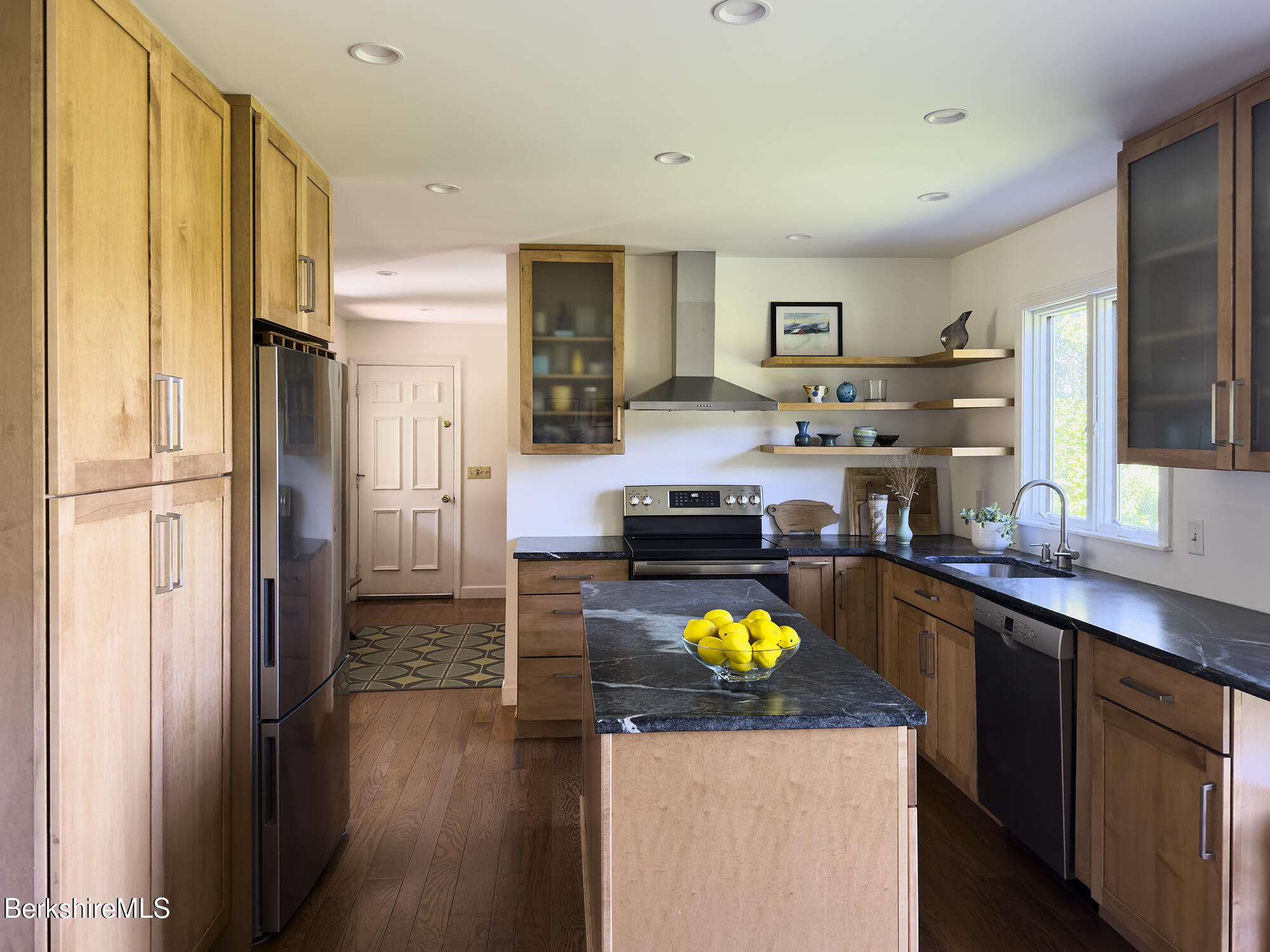 Kitchen with a stone-topped island, light wood cabinets, and open shelves.