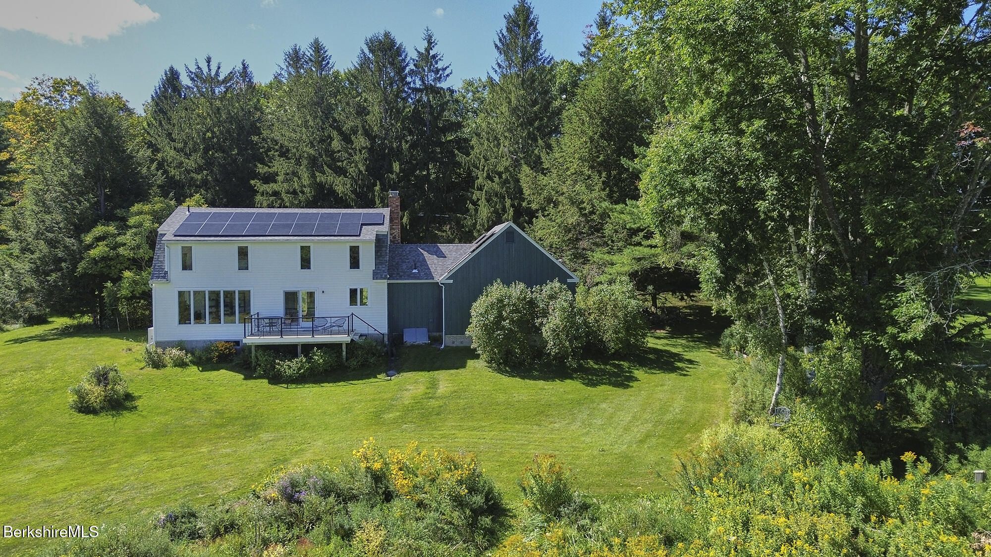 White home with rooftop solar panels and a deck, surrounded by trees.