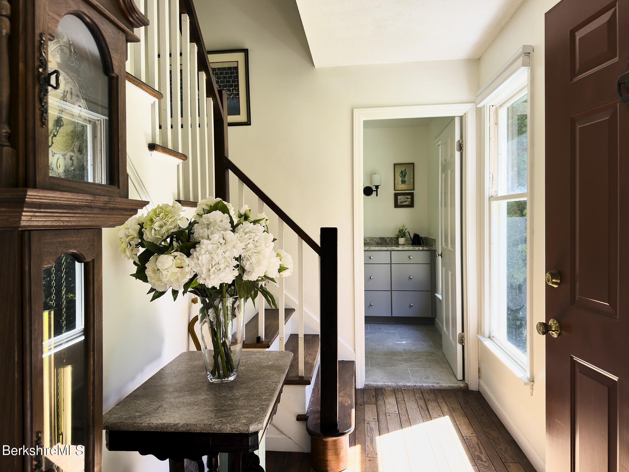 Entry hall with a dark wood staircase, grandfather clock, and white flowers on a table.