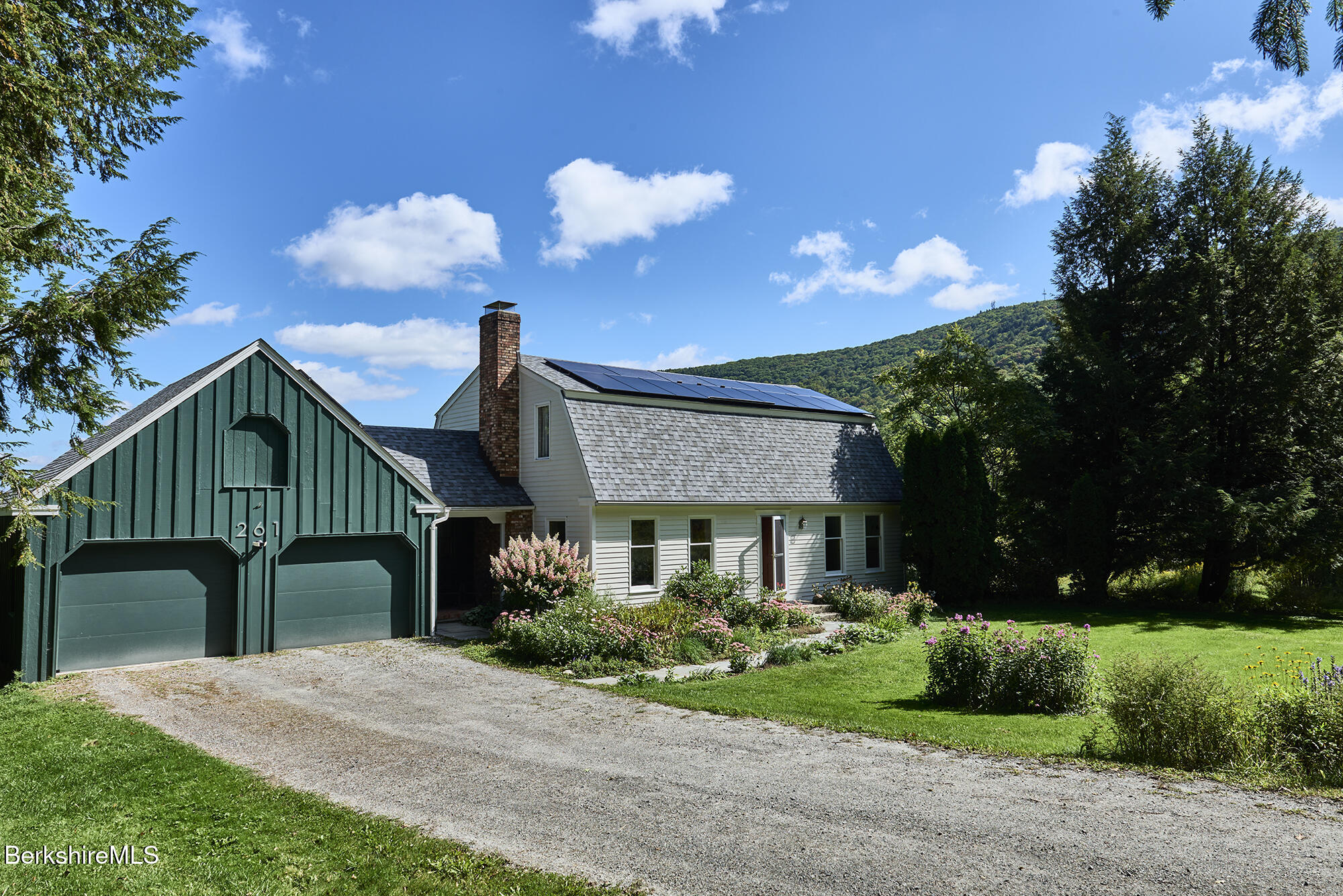 House with dark green two-car garage, rooftop solar panels, flower gardens, and a wooded hillside.