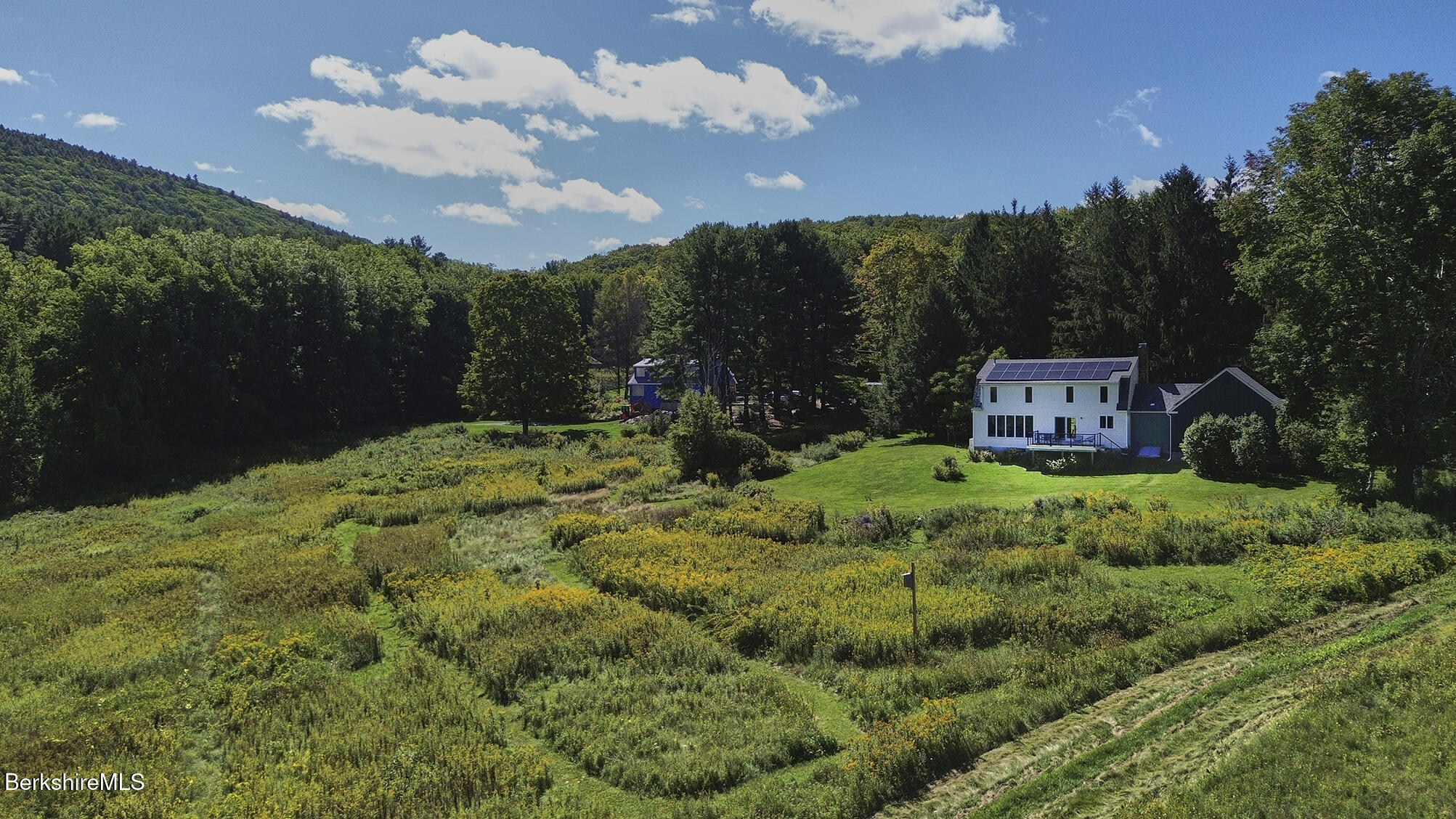 White house with rooftop solar panels sits in a meadow bordered by woods and rolling hills.