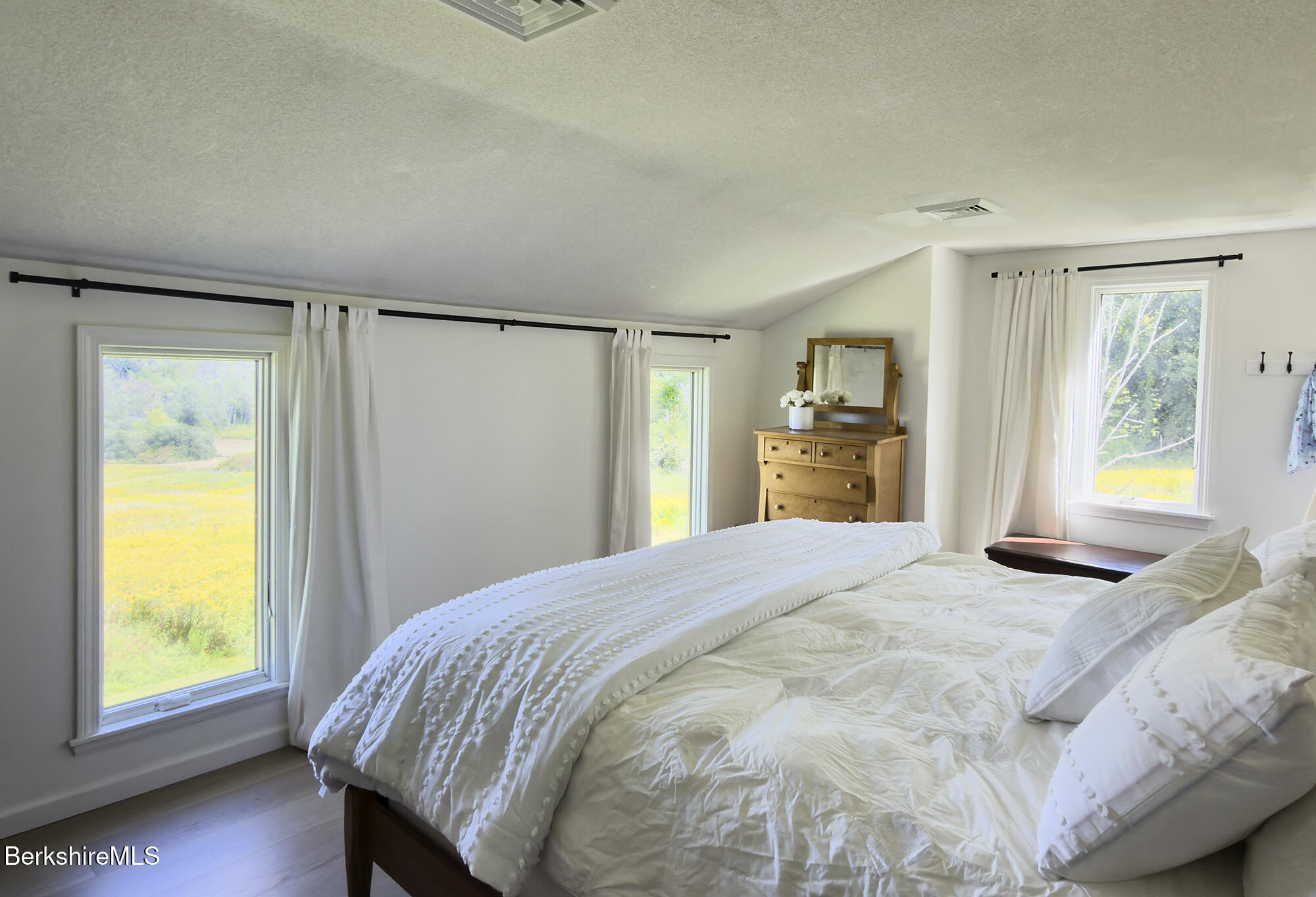 Bedroom with a white bed, three curtained windows, and a wooden dresser with mirror.
