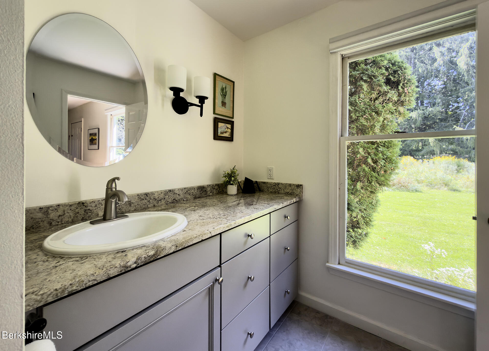 Bathroom vanity with a round mirror, stone countertop, and window overlooking a green lawn.