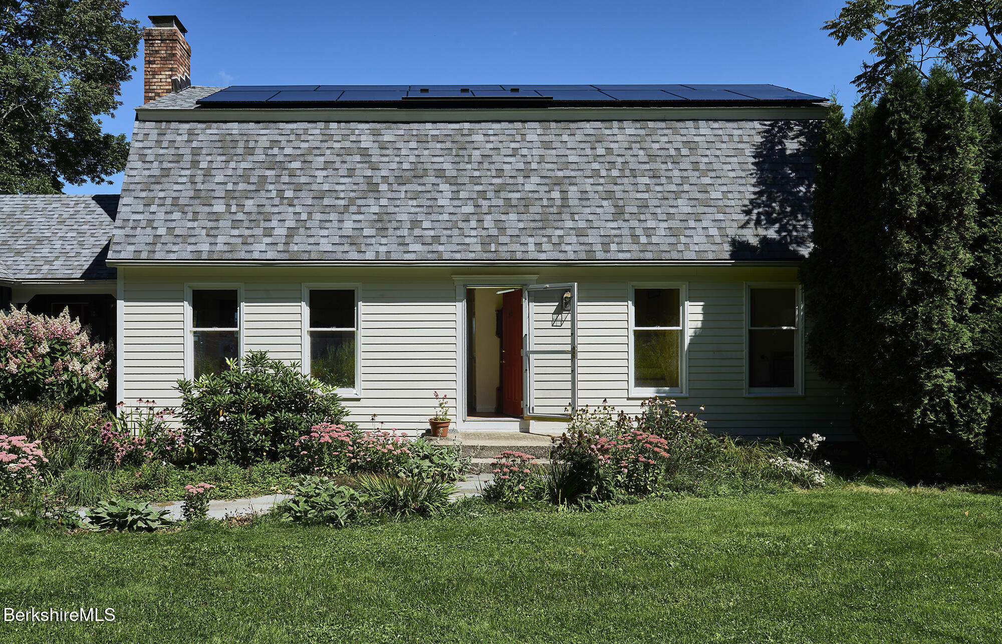 House with rooftop solar panels, an open front door, and flowering garden beds.