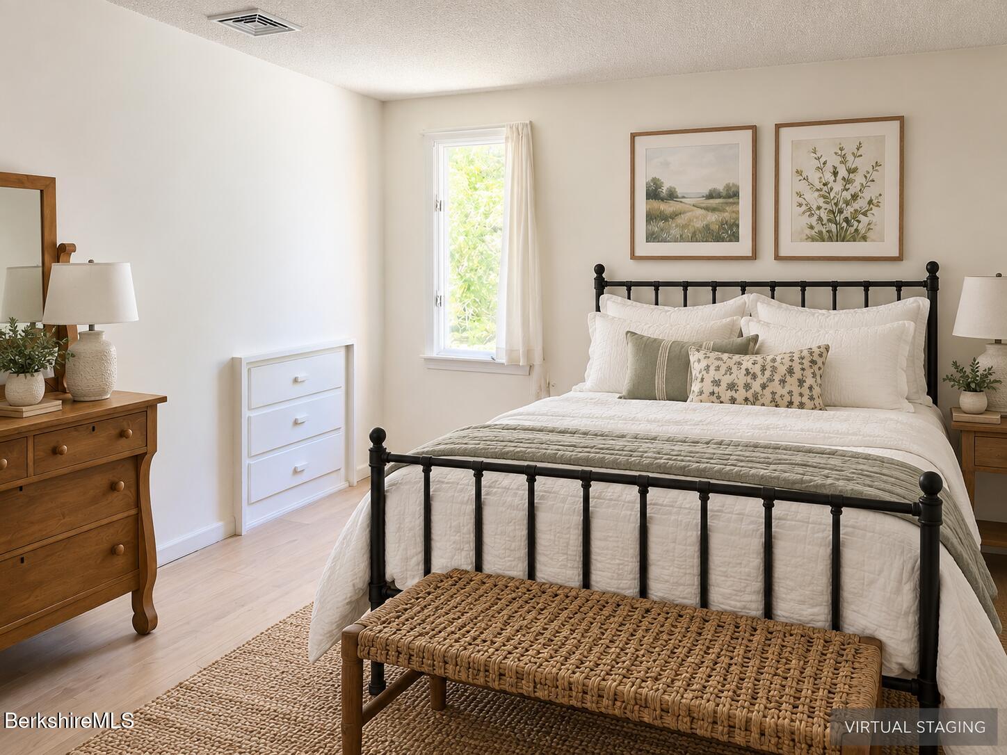Bedroom with a black metal bed, white bedding, botanical artwork, and a woven bench.