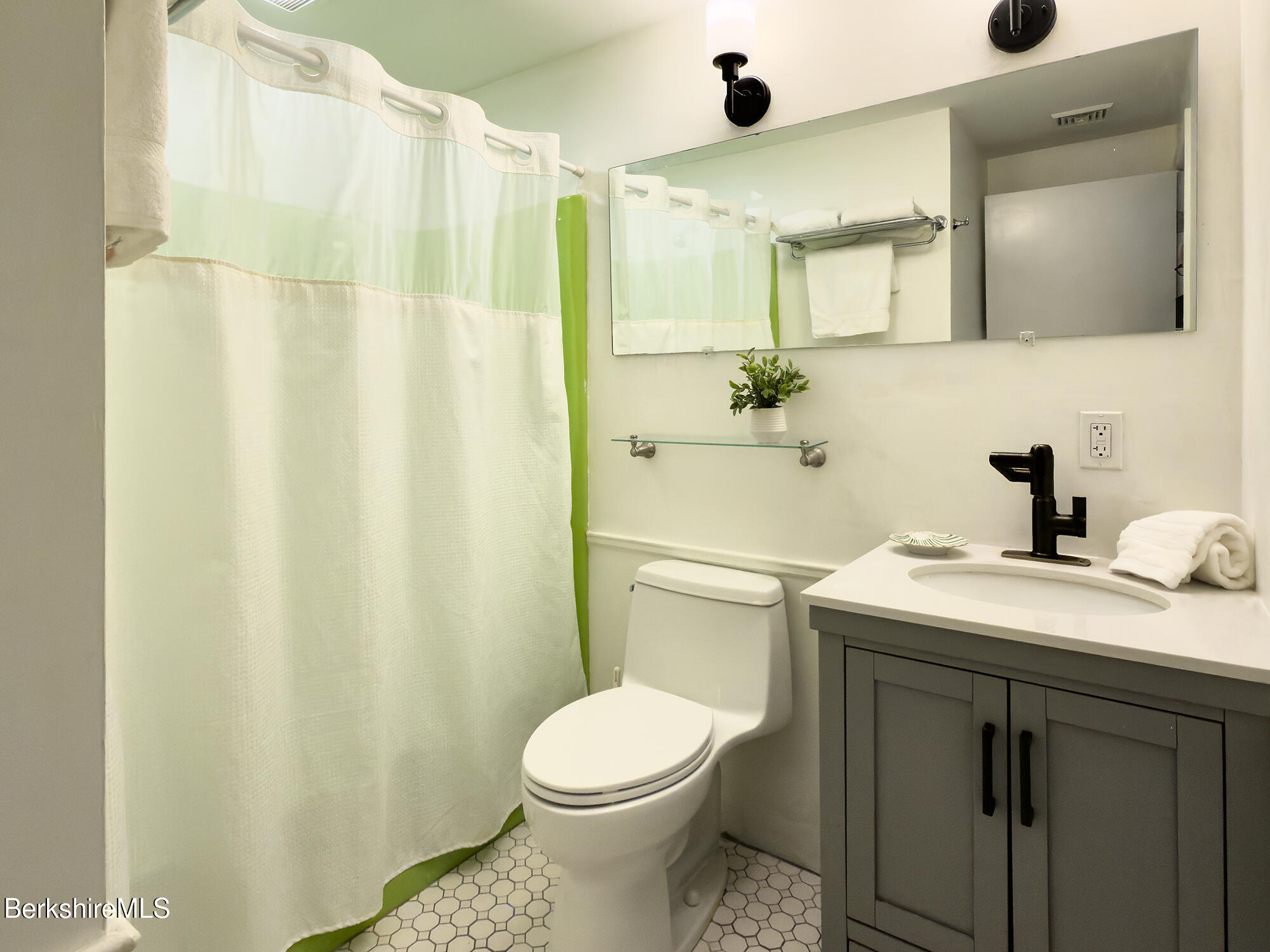 Bathroom with a green-edged shower curtain, gray vanity, black faucet, and white hexagonal floor tile.