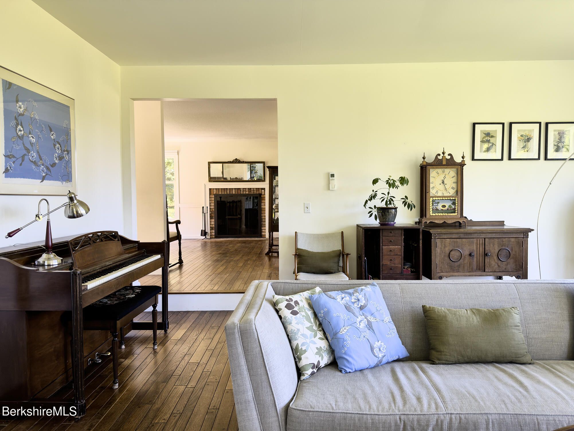Living room with a gray sofa, upright piano, hardwood floors, and an adjoining room with a brick fireplace.