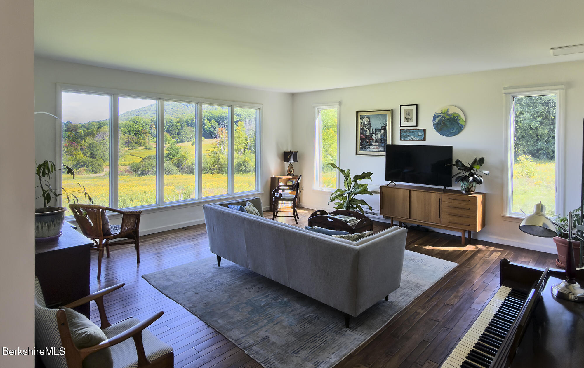 Living room with broad meadow-facing windows, a gray sofa, and a piano.