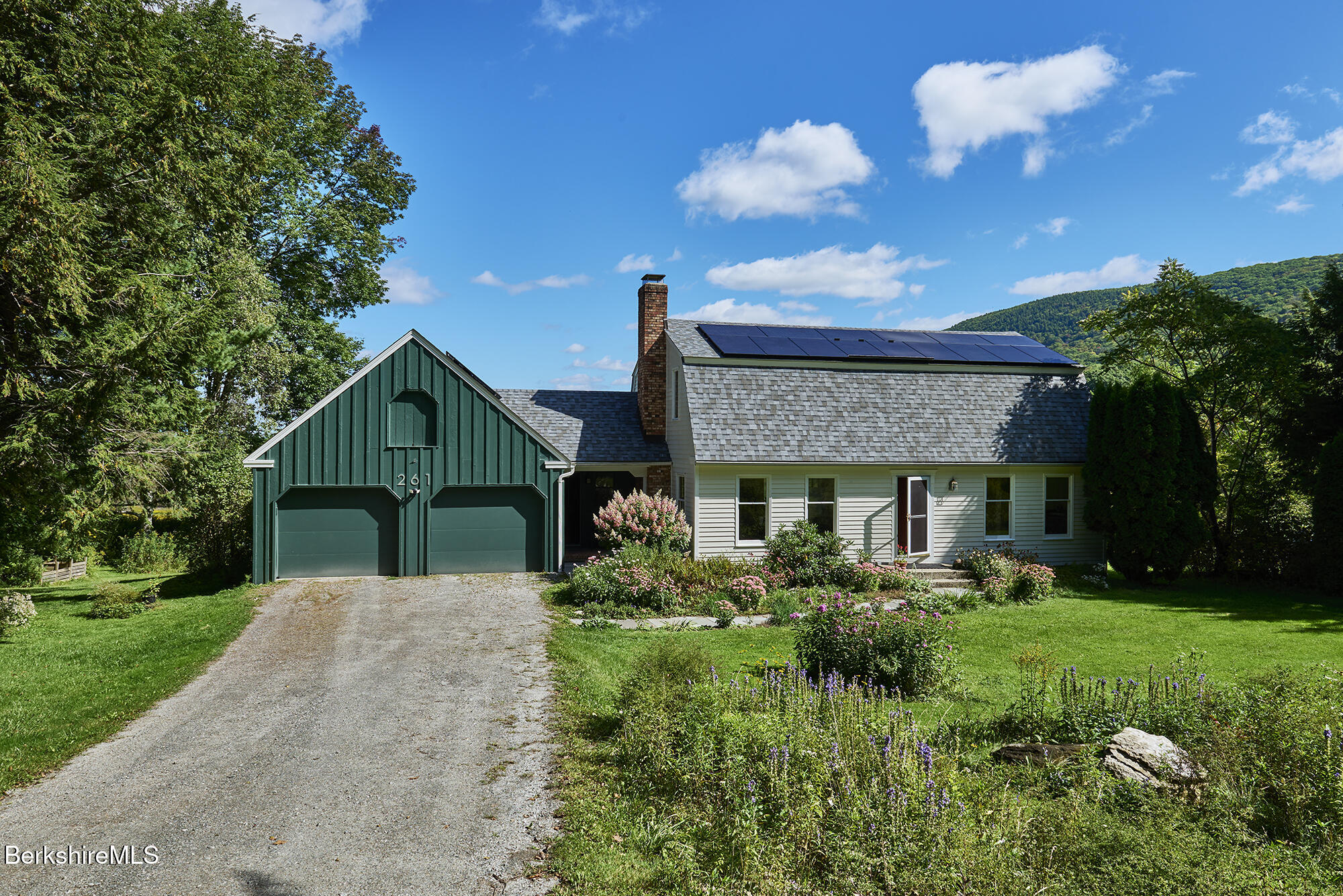 Home with a dark green two-car garage, rooftop solar panels, and flowering gardens beside a gravel driveway.