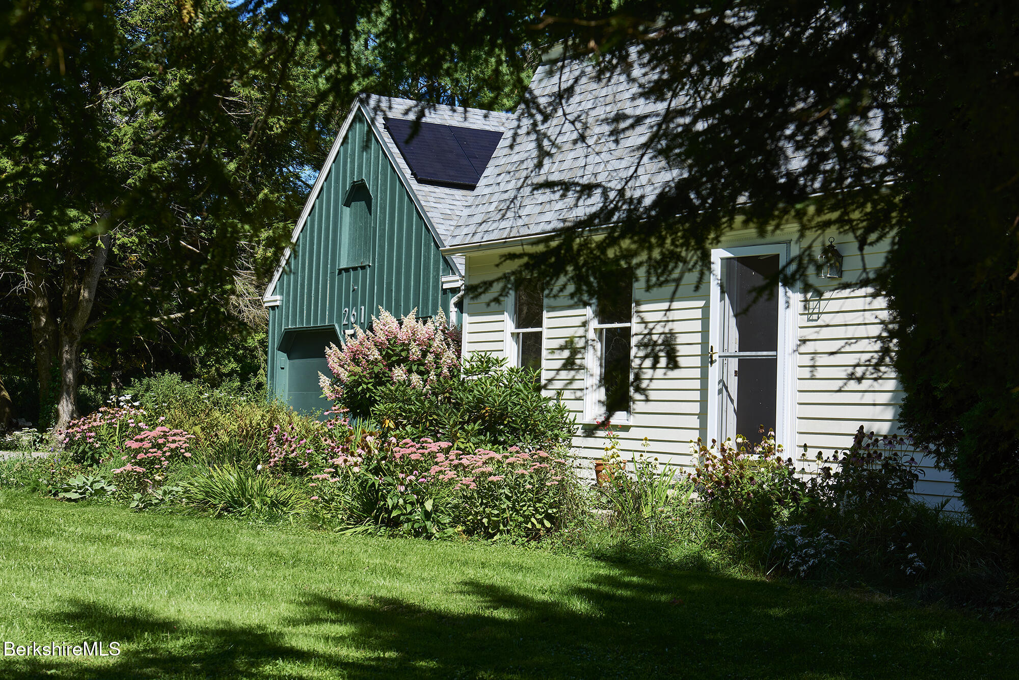 House with a teal gable, solar panels, and flowering garden beneath mature trees.
