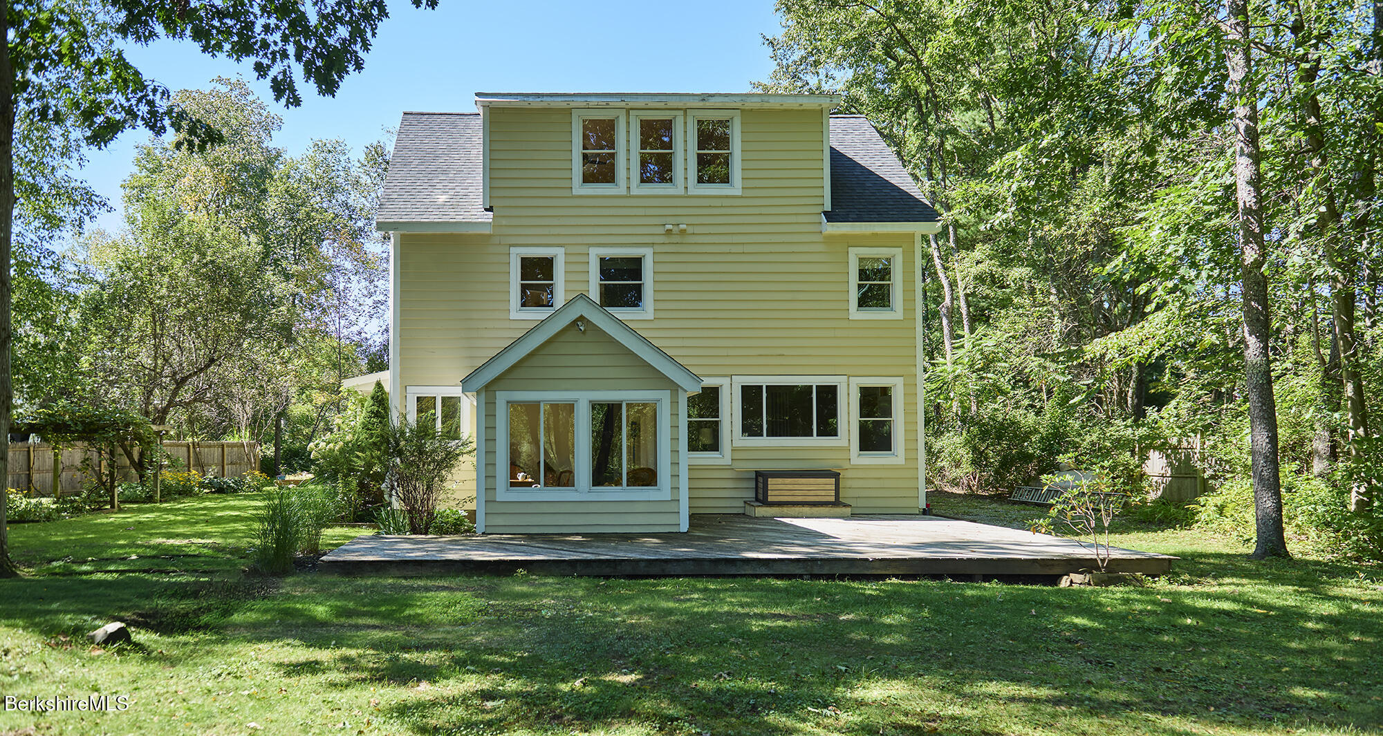 Rear of a yellow-sided house with an enclosed porch and broad deck, surrounded by trees.