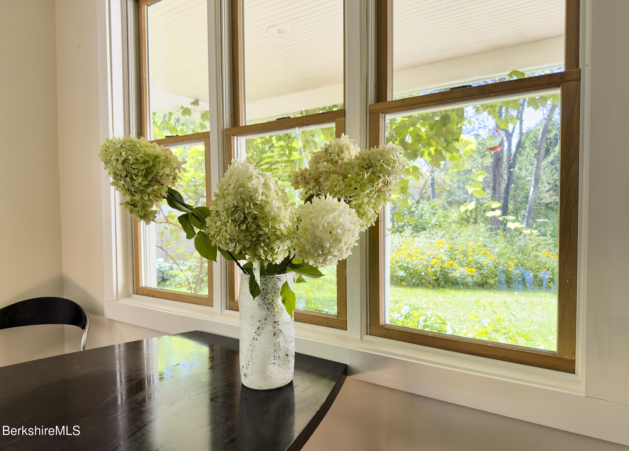 Dining area with white hydrangeas on a dark table beside large garden-facing windows.