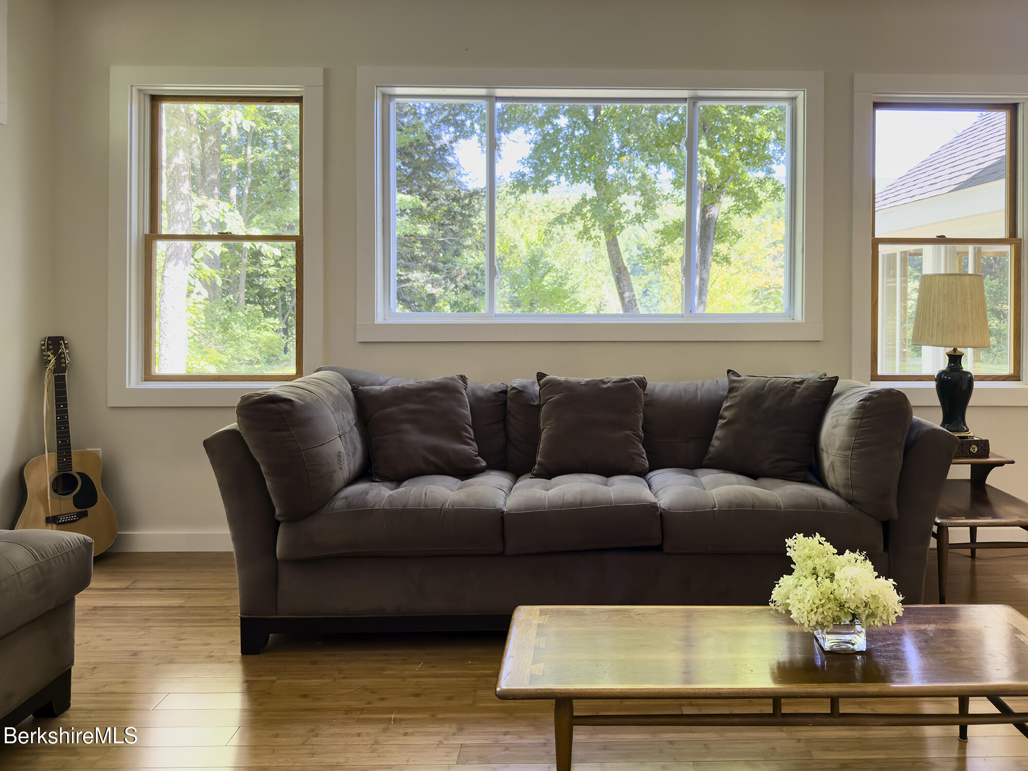 Living room with a dark sofa beneath large windows overlooking leafy trees, with a guitar and wooden coffee table.