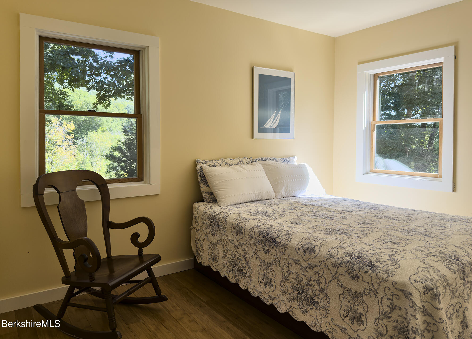 Bedroom with a patterned bedspread, antique rocking chair, and two windows overlooking trees.