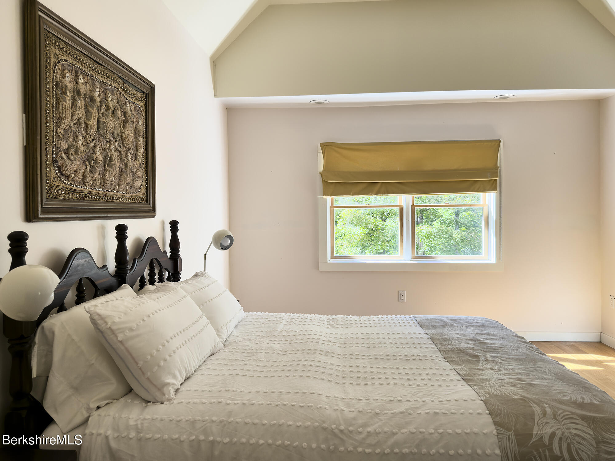 Bedroom with a textured white bed, carved dark headboard, framed artwork, and shaded window.
