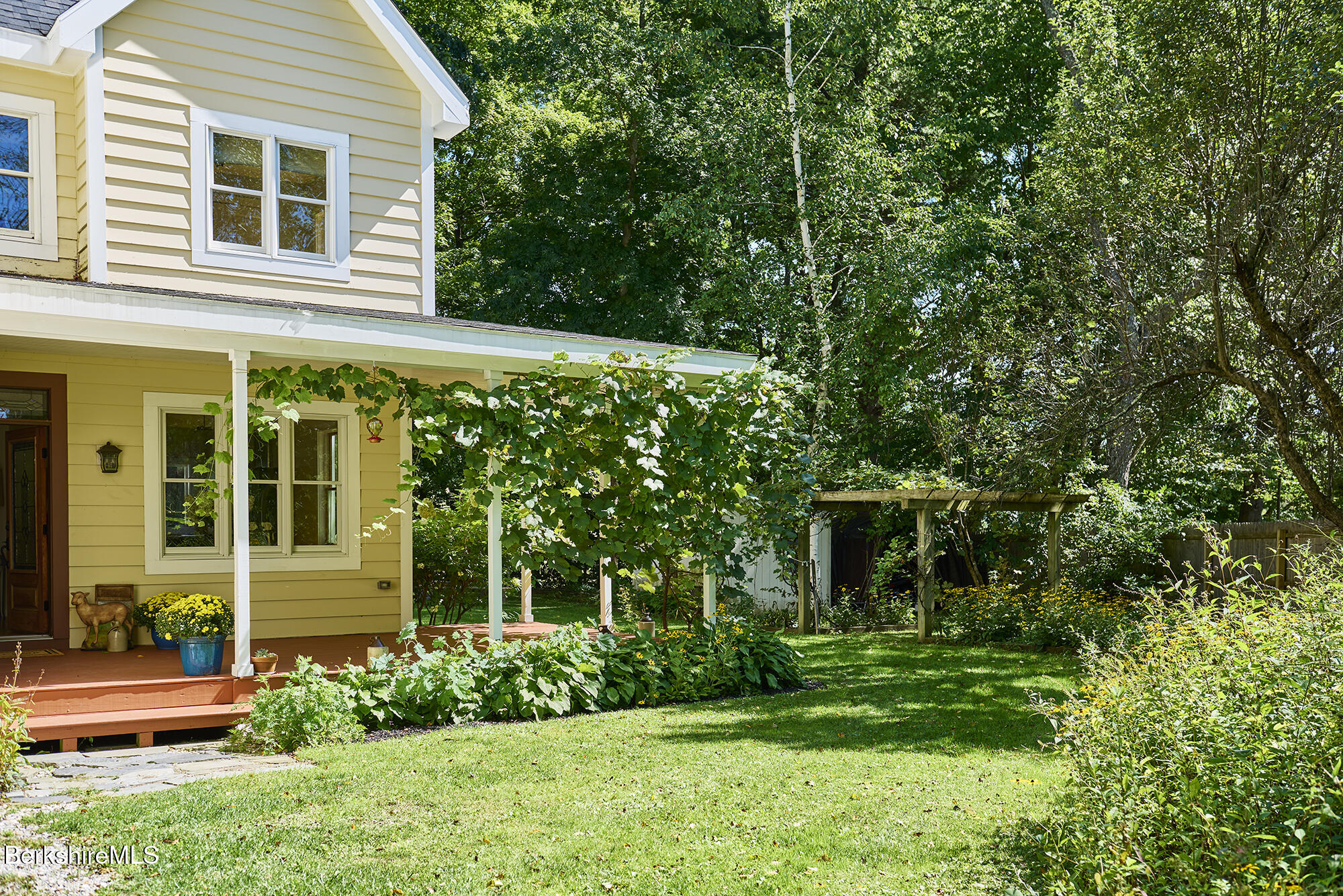Yellow clapboard house with a vine-covered porch, garden beds, and a wooden pergola beside the lawn.