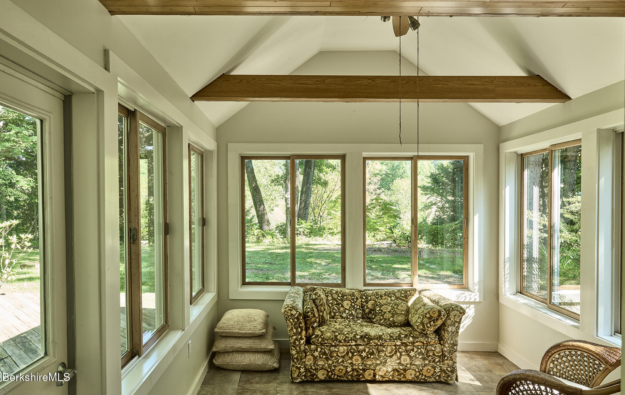 Sunroom with a floral sofa, exposed wood beams, and large windows overlooking a leafy yard.