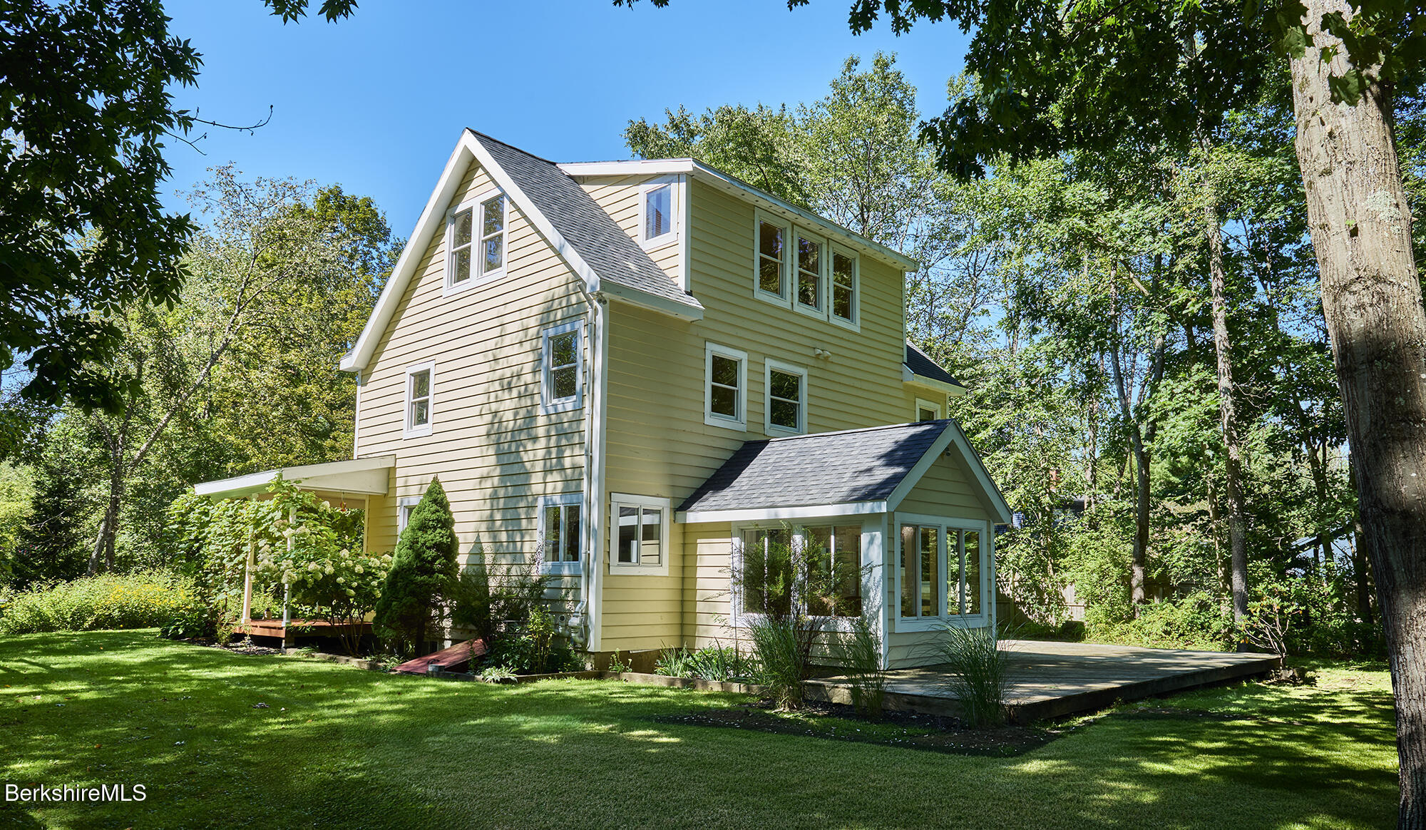 Pale yellow multi-story house with a glass-enclosed sunroom and wooden deck, surrounded by trees.