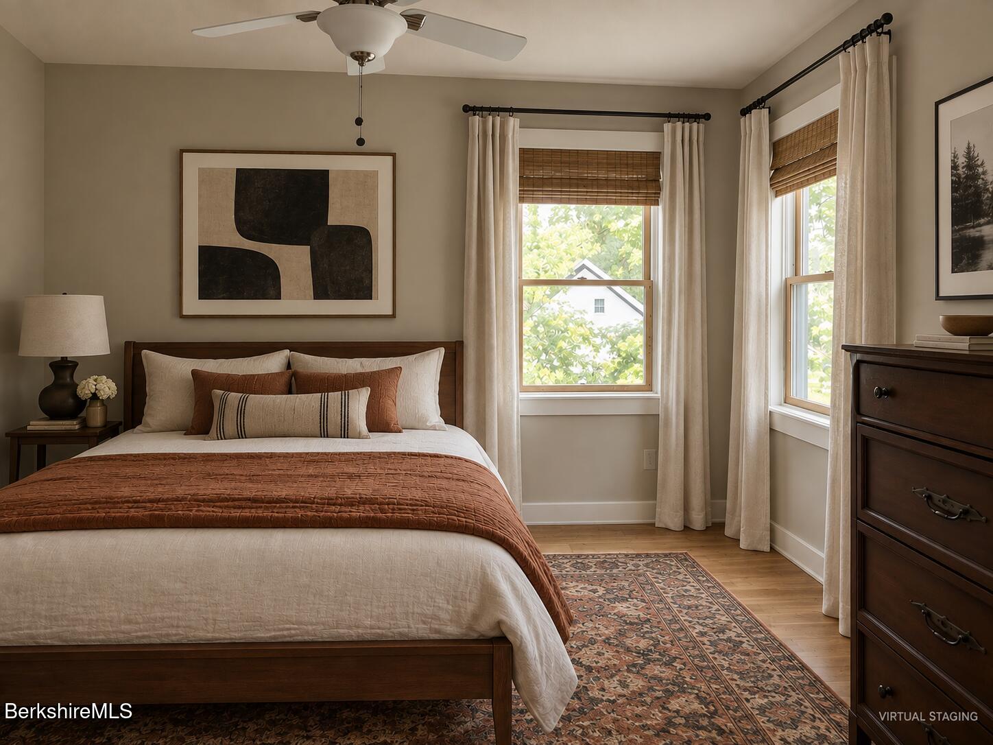 Virtually staged bedroom with a dark wood bed, rust-colored coverlet, patterned rug, and two bright windows.