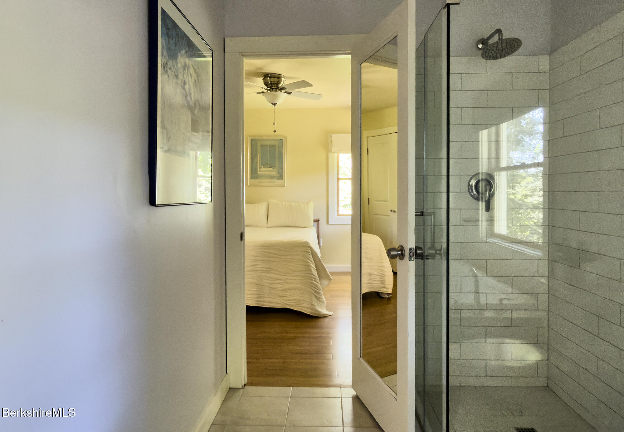 Bathroom with glass subway-tile shower opening toward a bedroom with a bed and hardwood floor.