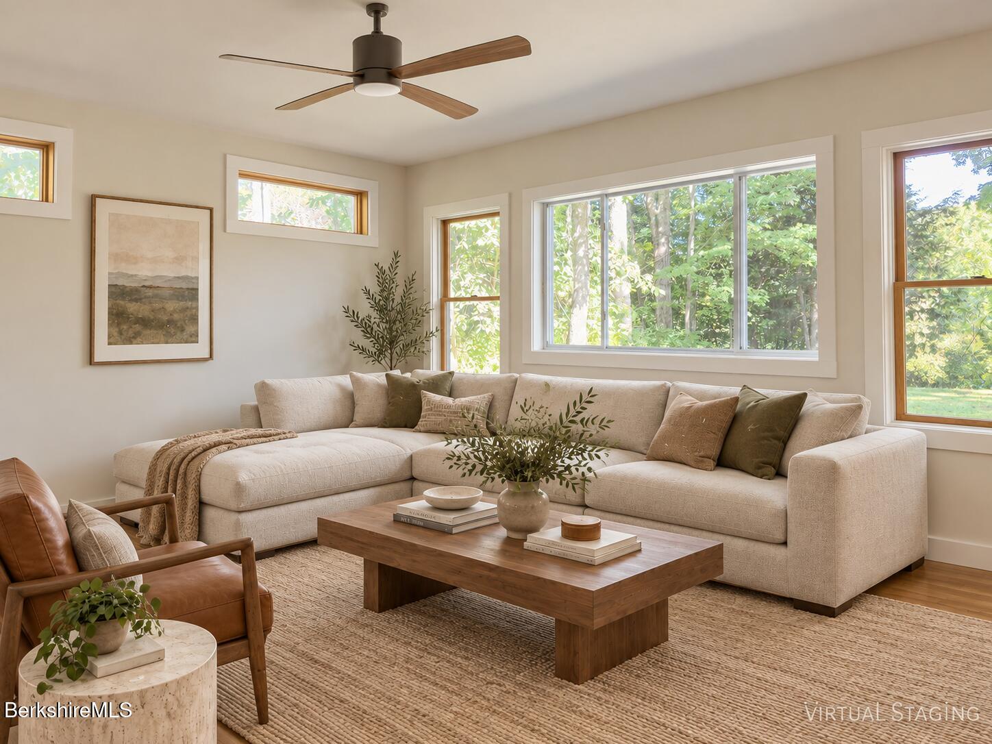 Virtually staged living room with a cream sectional, wood coffee table, and wide windows overlooking greenery.
