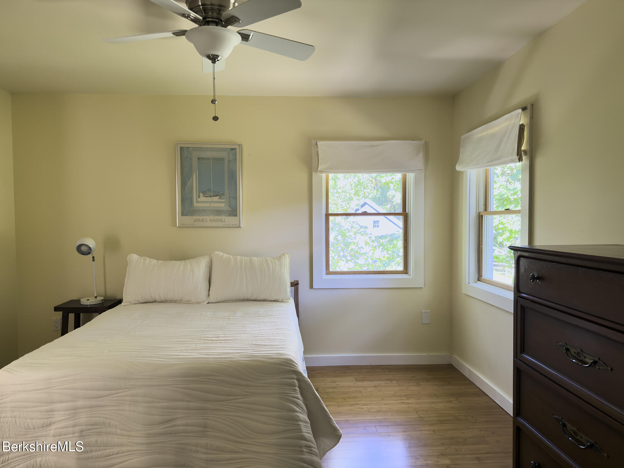 Bedroom with a white bed, two bright windows, hardwood floors, and a dark wood dresser.