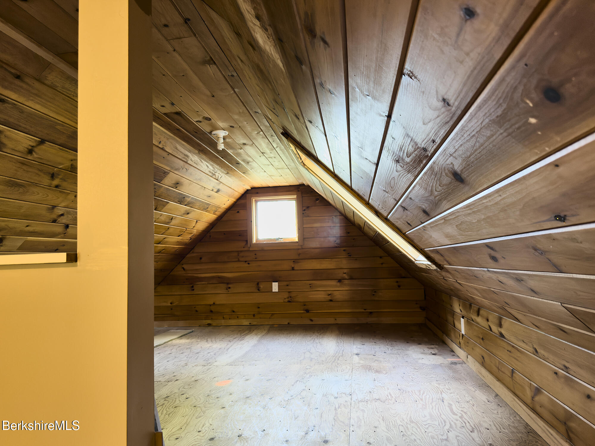 Unfinished attic with sloped wood-plank walls, plywood flooring, and a small window.