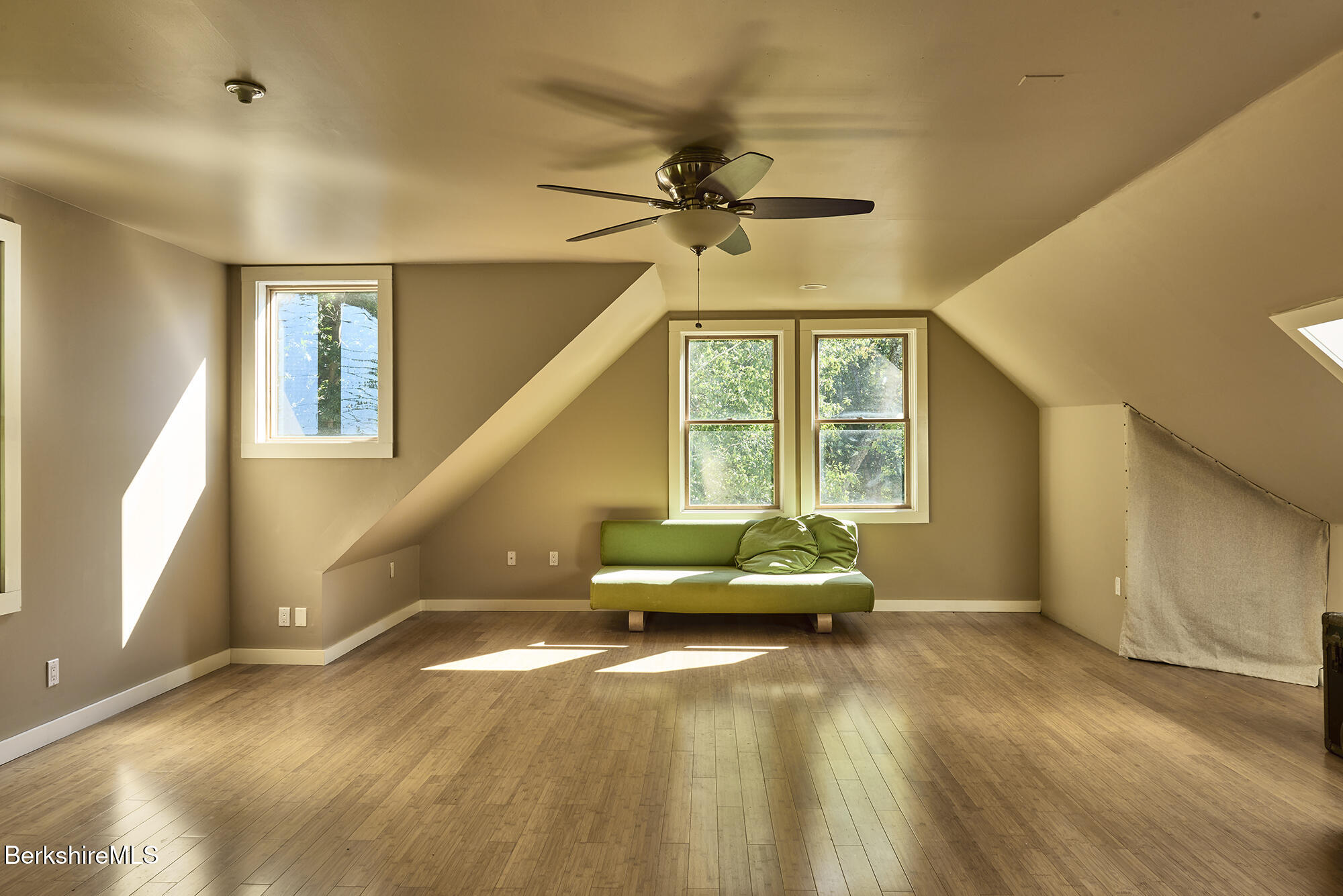 Finished attic room with sloped ceilings, wood flooring, green loveseat, and windows overlooking trees.