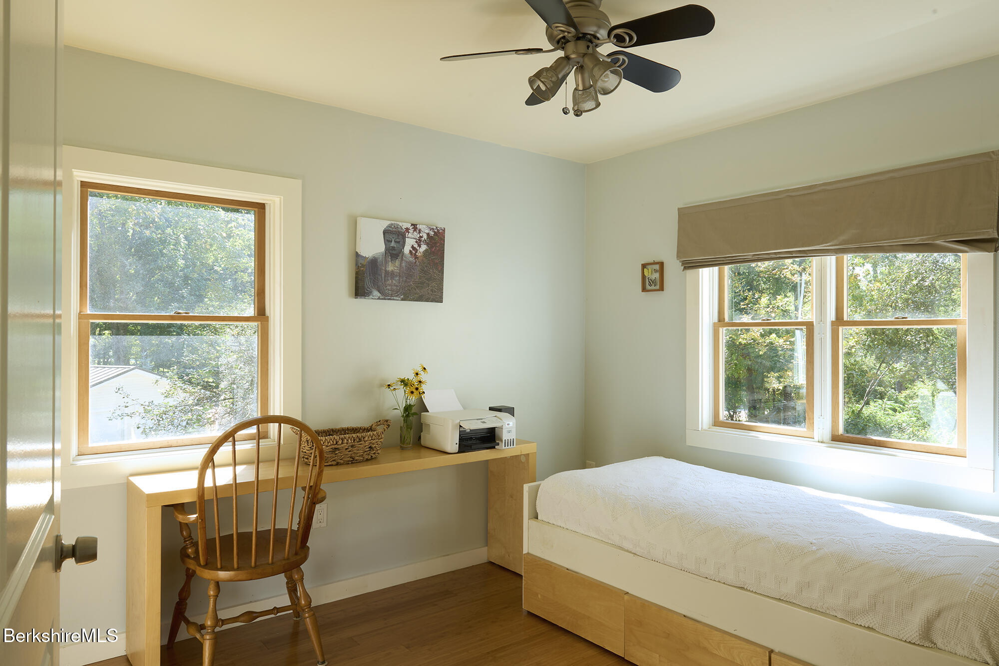 Bedroom with a bed, wooden desk, and large windows overlooking leafy trees.