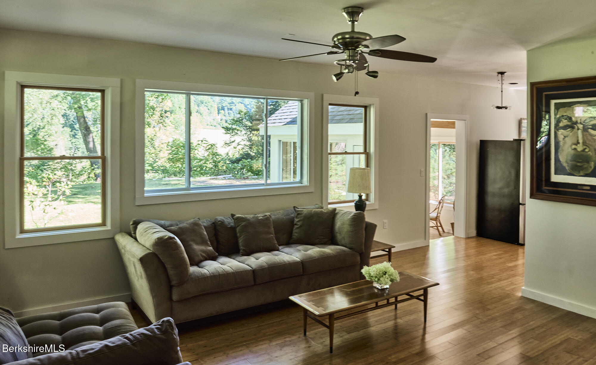 Living room with a gray sofa, hardwood floors, and large windows overlooking a leafy yard.