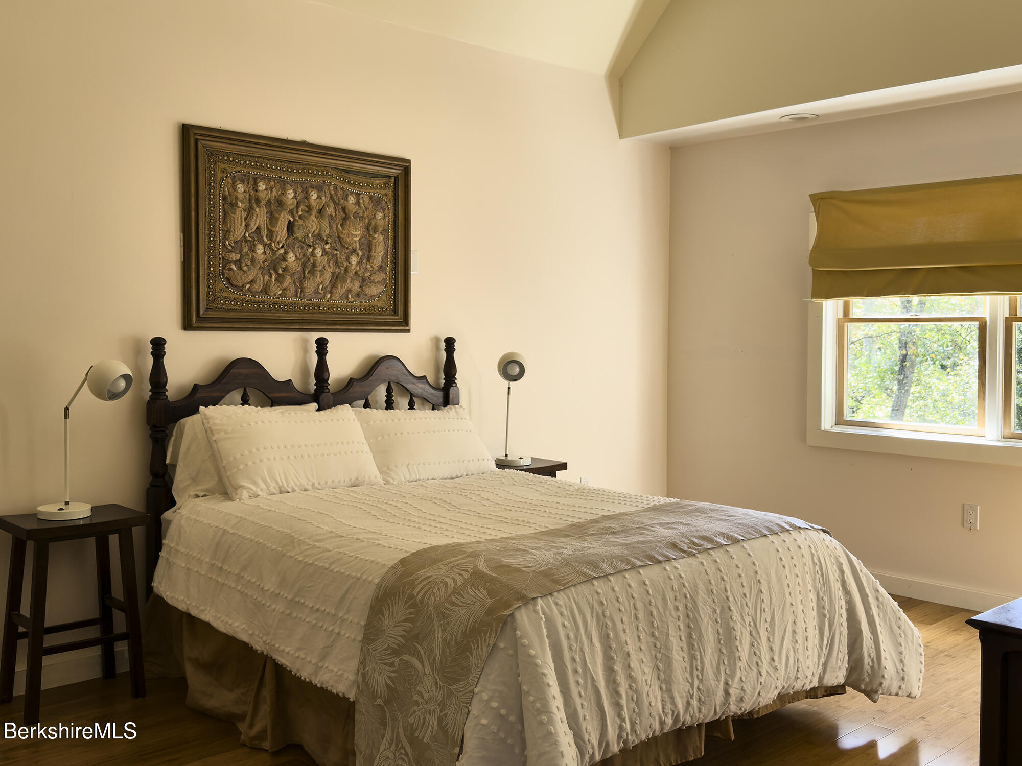 Bedroom with a carved dark wood bed, cream bedding, framed artwork, and a shaded window.