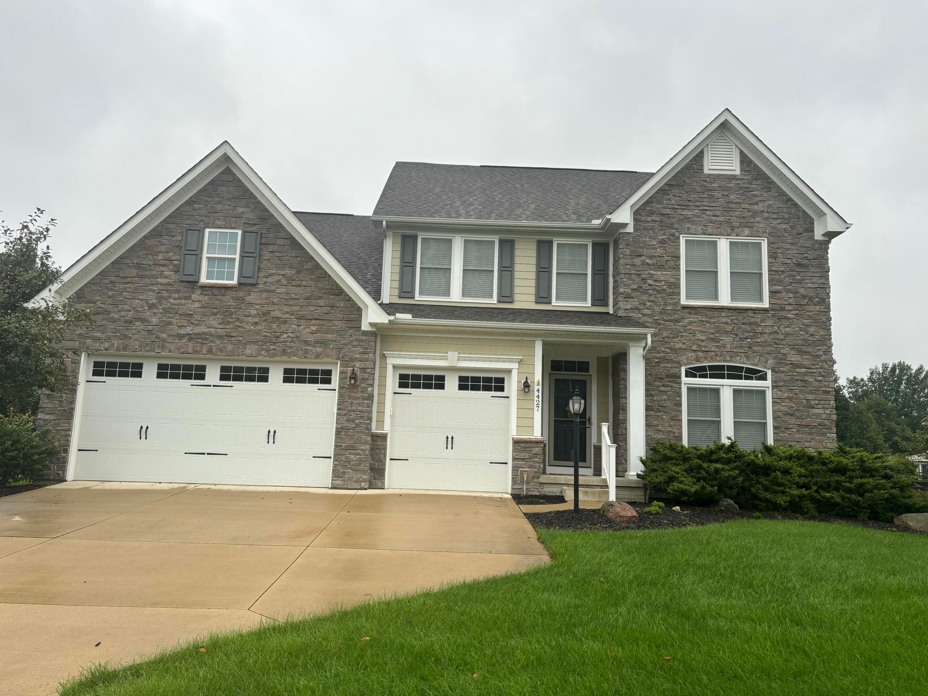 Two-story stone-front home with two white garage doors, a wide driveway, and a green lawn.