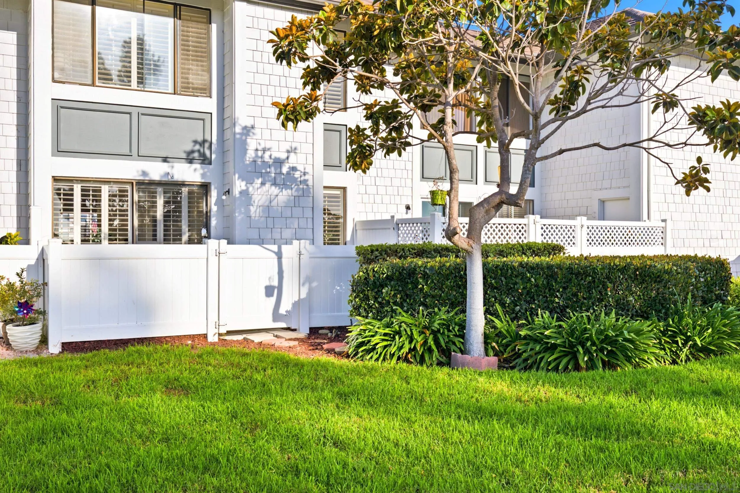 White shingle-sided home behind a fence, with a green lawn and mature tree.