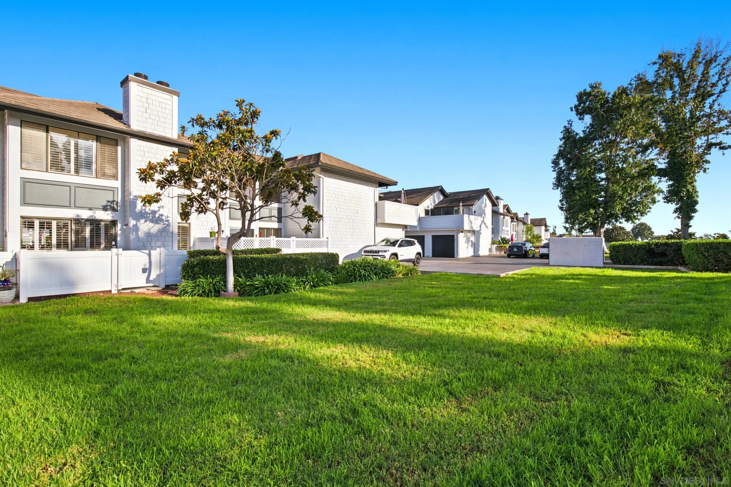 White two-story townhouse beside a broad green lawn, with neighboring homes and parked cars.